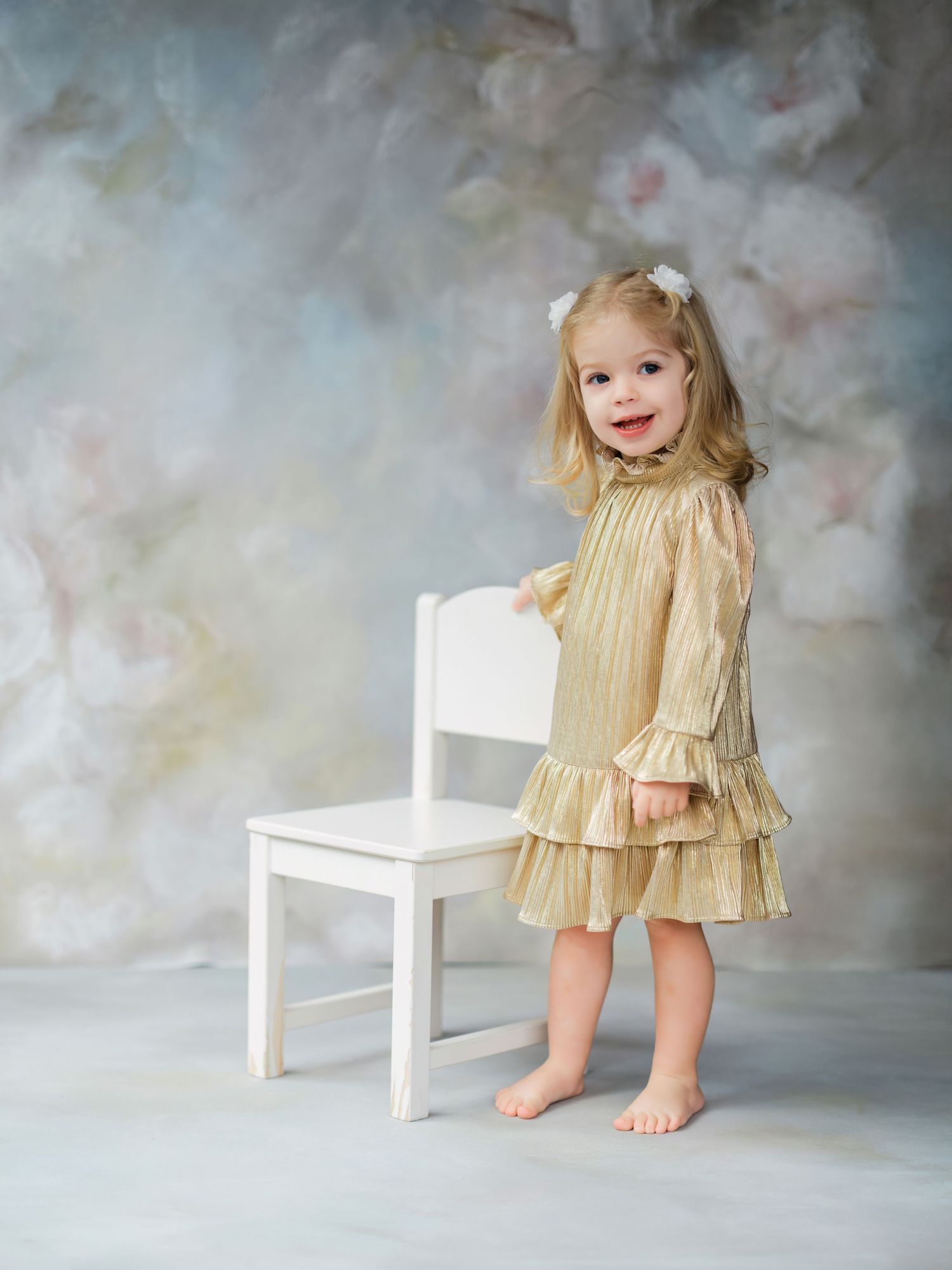 Young child in a gold ruffled dress stands barefoot next to a white wooden chair against a cloudy gray backdrop.