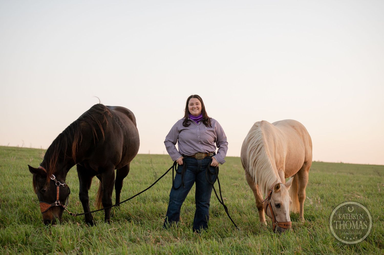 A person stands confidently between two grazing horses in a sunlit pasture.