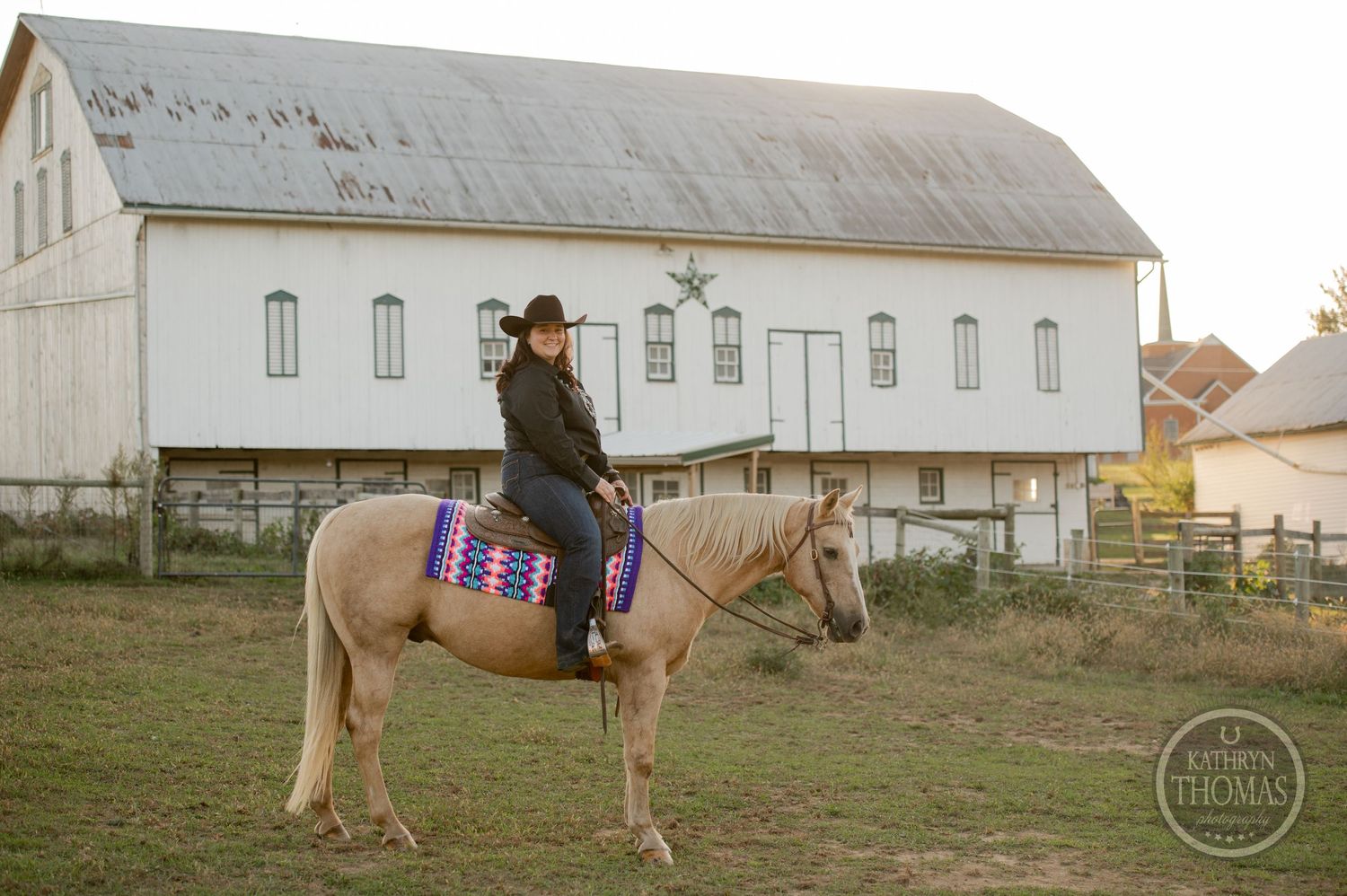 Pale horse with colorful saddle blanket stands in front of white barn building.