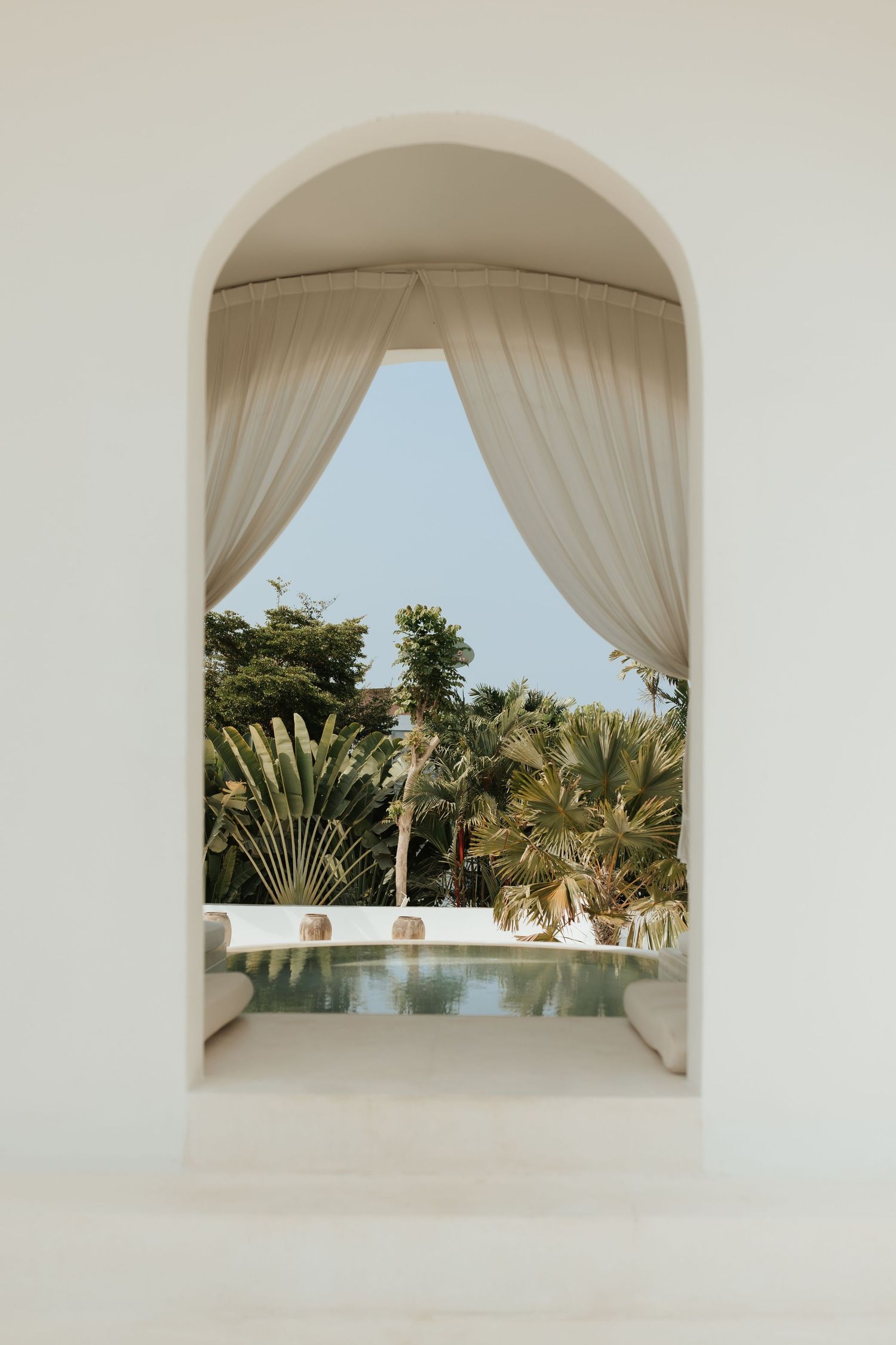 View through white arched doorway with curtains revealing tropical garden and swimming pool surrounded by palm trees.