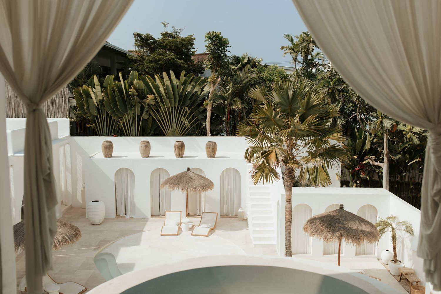 Tropical courtyard with white stucco walls, palm trees, and natural woven umbrellas around a curved pool.