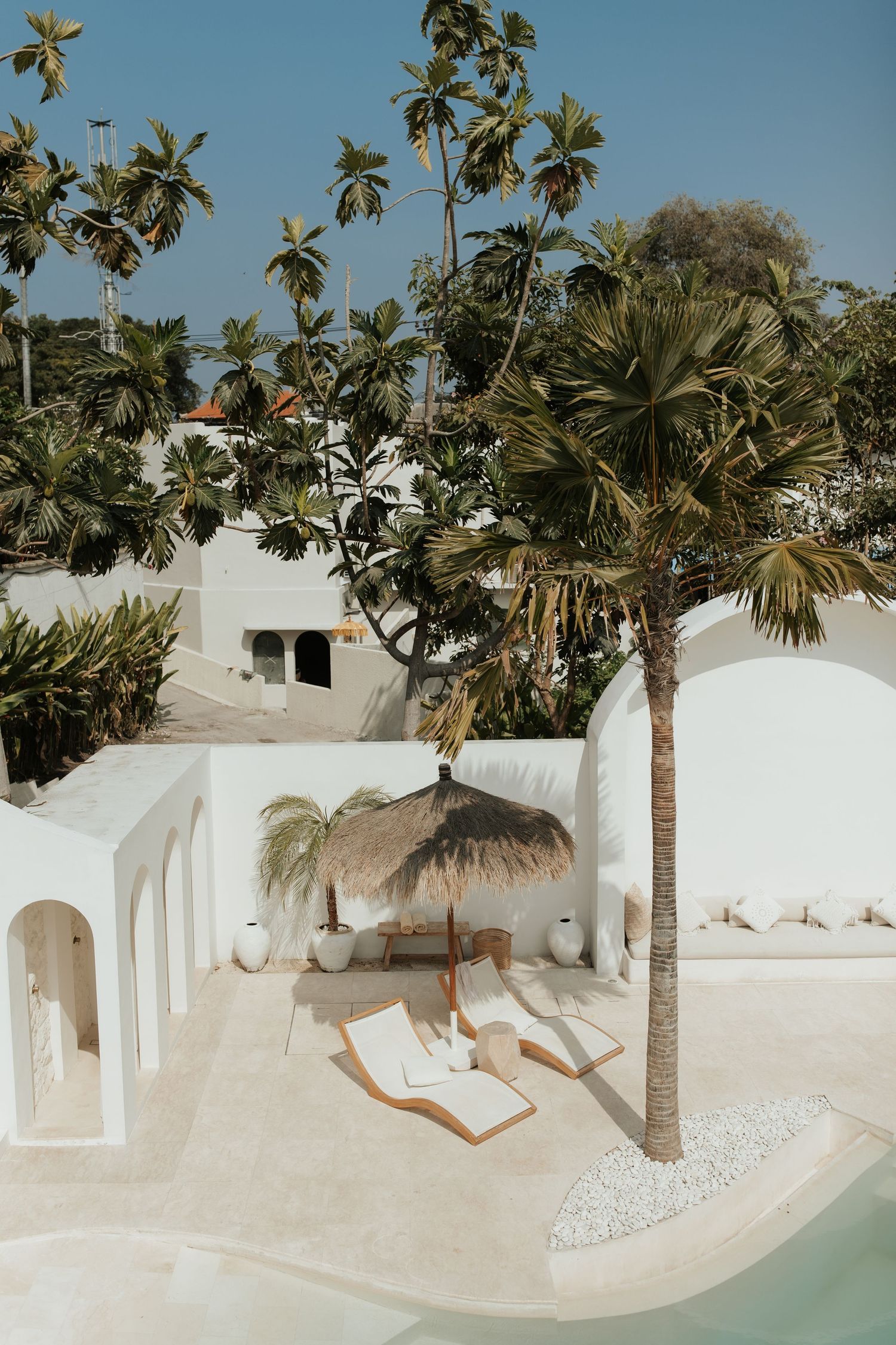 Luxurious white Mediterranean resort with palm trees and lounge chairs beside a turquoise pool on a sunny day.