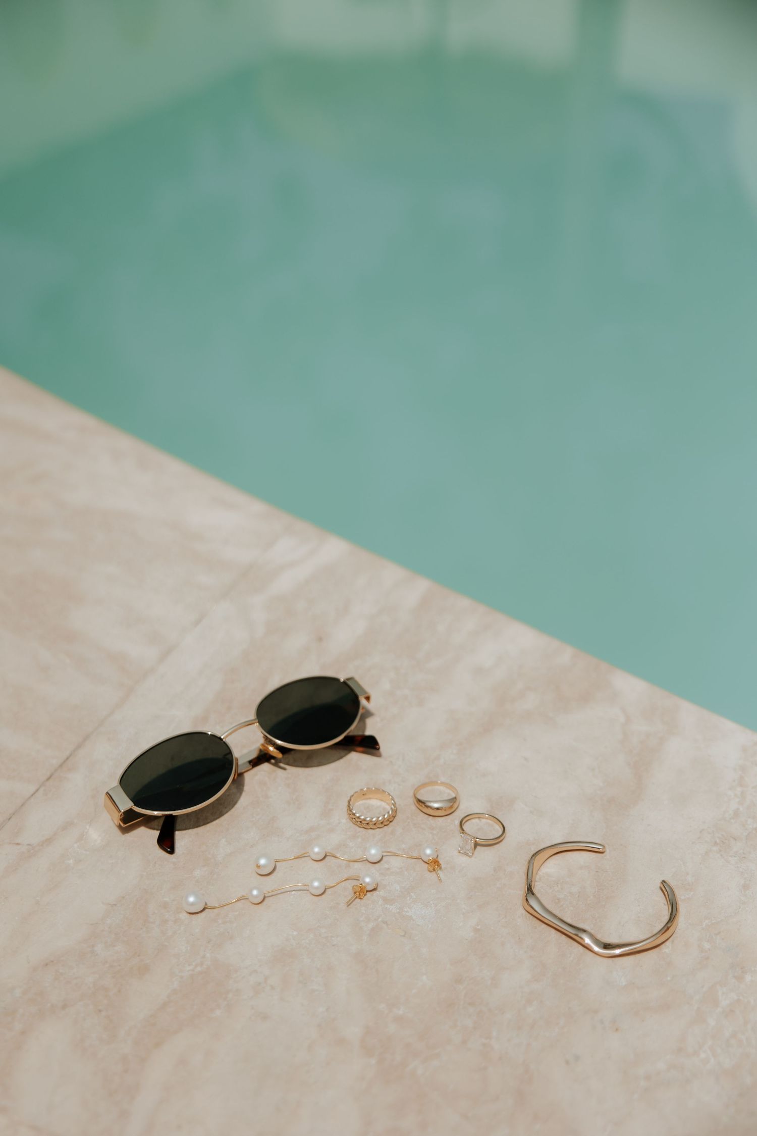 Sunglasses and gold jewelry including rings and a bracelet arranged on a beige surface next to turquoise water.