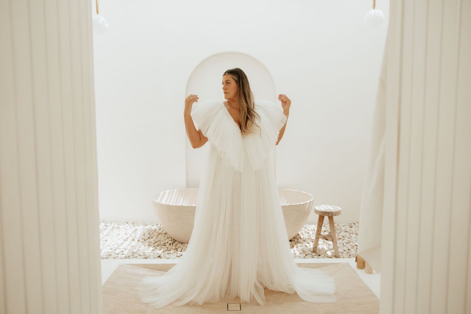 A bride in a flowing white dress stands between sheer curtains in a bright sunlit room during her wedding preparations.