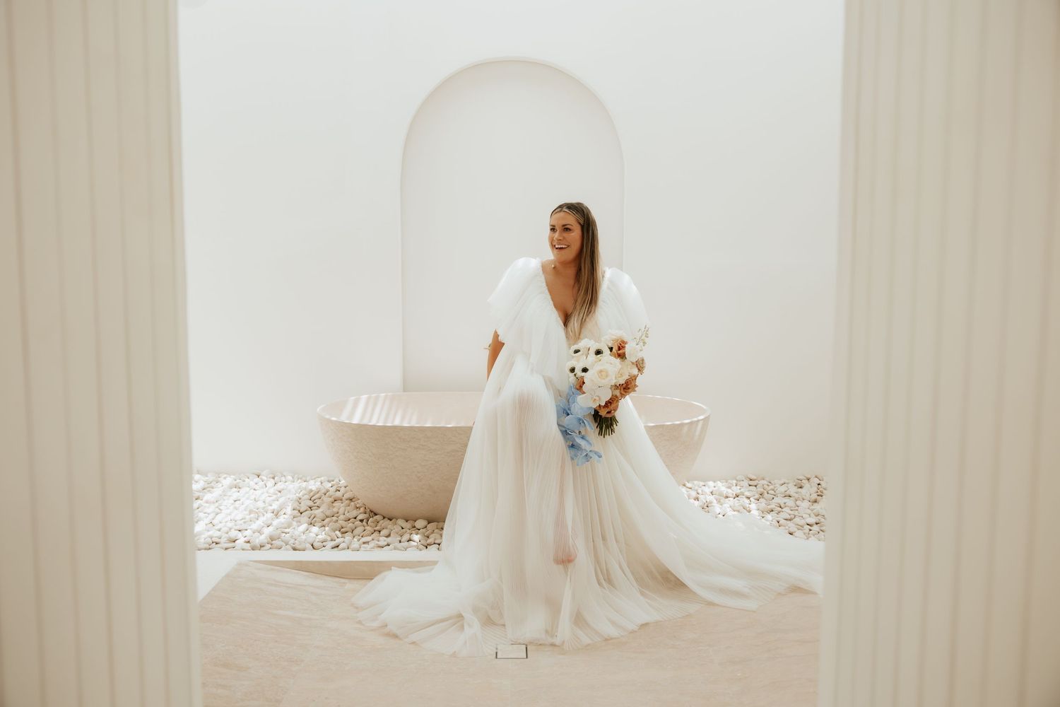 An elegant bride in a flowing white dress stands under an archway holding a bouquet.