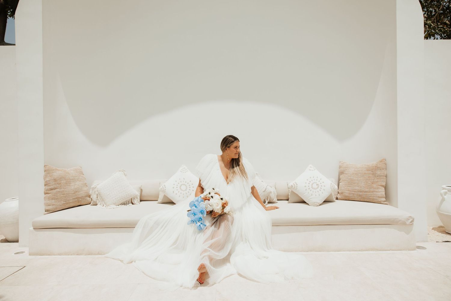 Person in white dress seated on cushioned bench in white arched alcove with pool view.