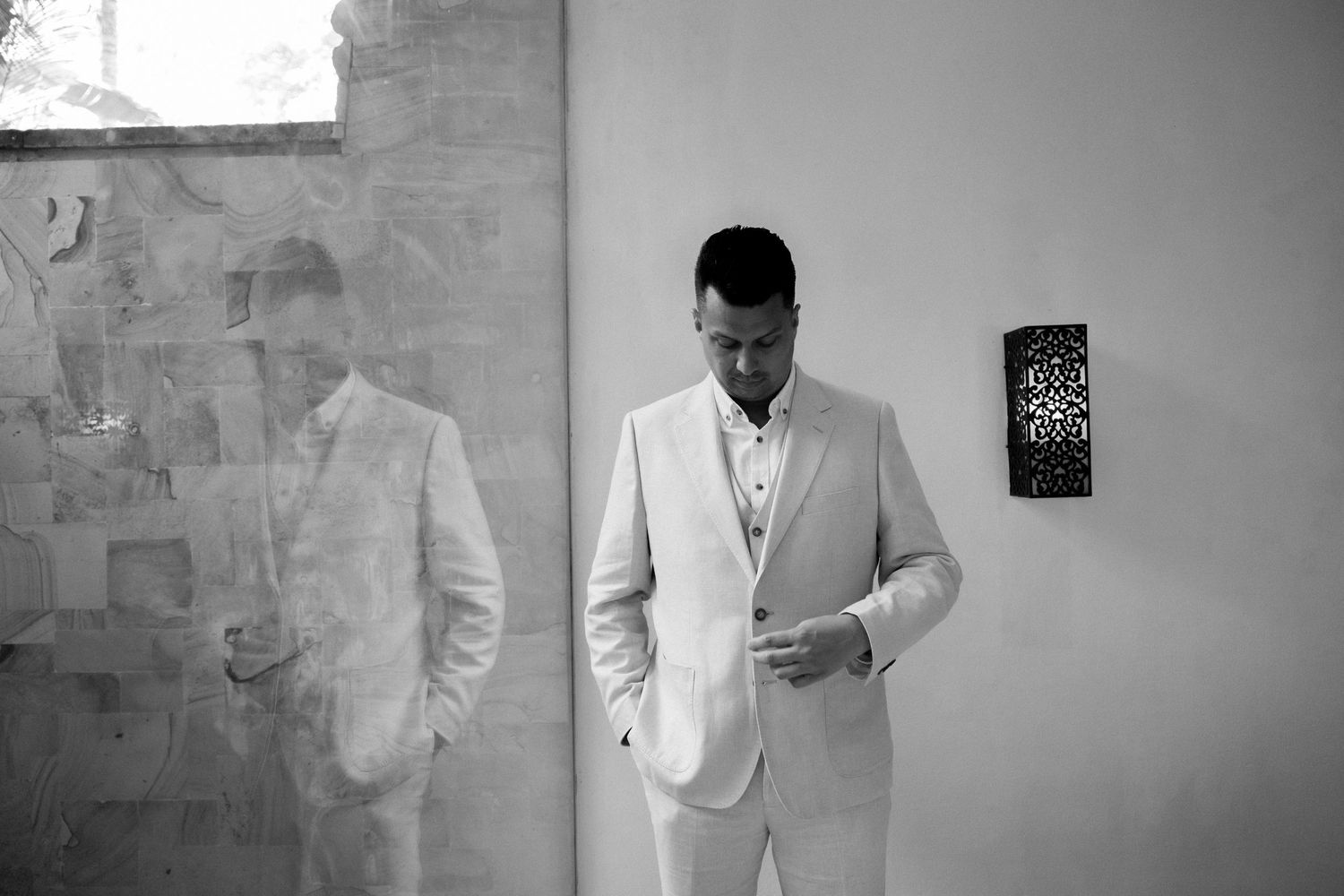 A person in a light colored suit jacket stands thoughtfully against a minimalist wall with a vintage black and white effect.