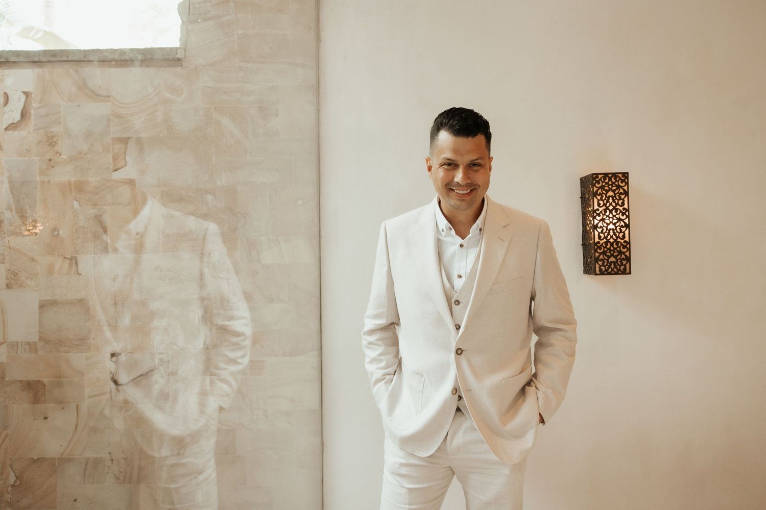 A person in a crisp white suit stands with hands in pockets against a textured beige wall with decorative sconce.