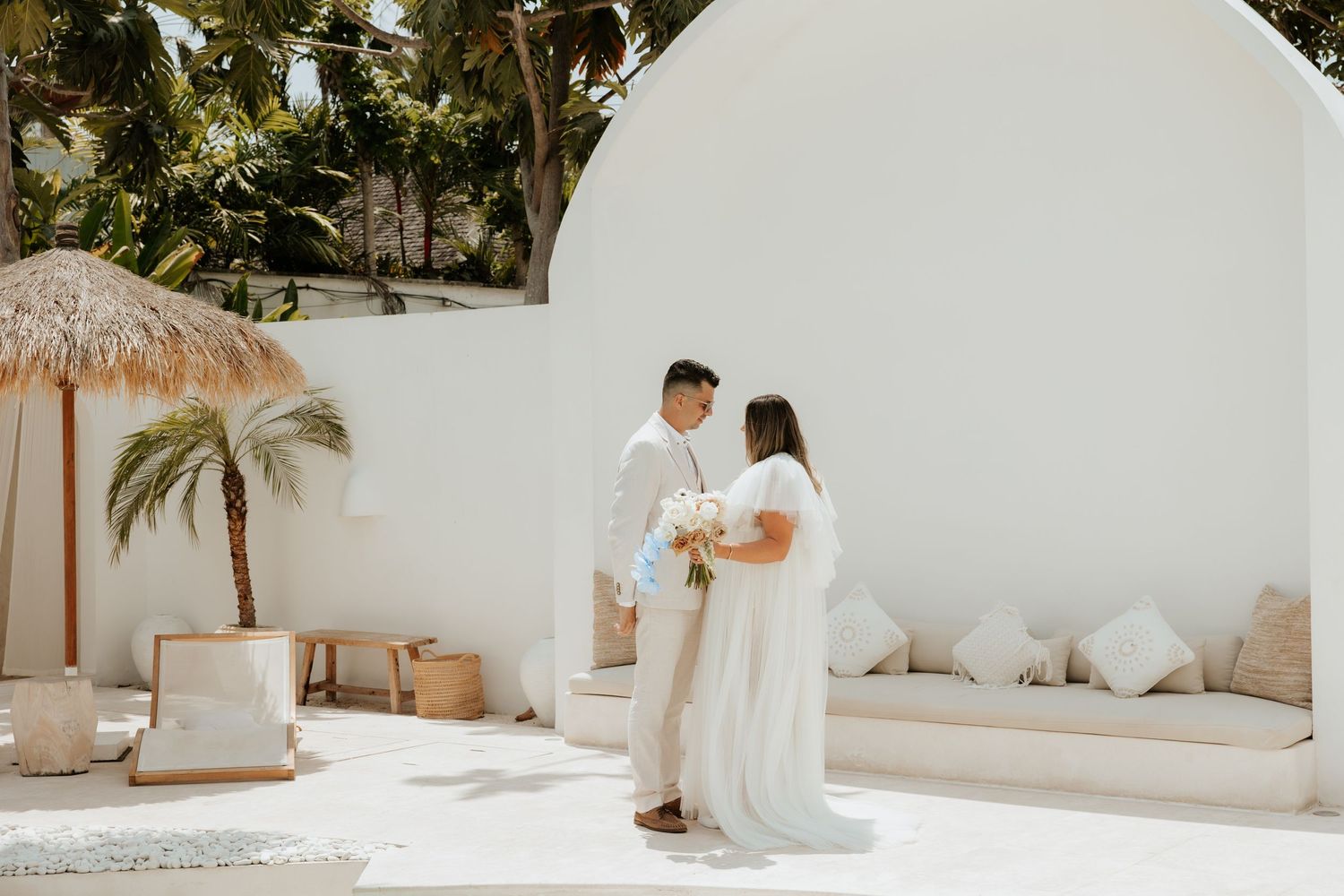 A couple stands intimately together in a sun-drenched courtyard with white walls and a thatched beach umbrella.