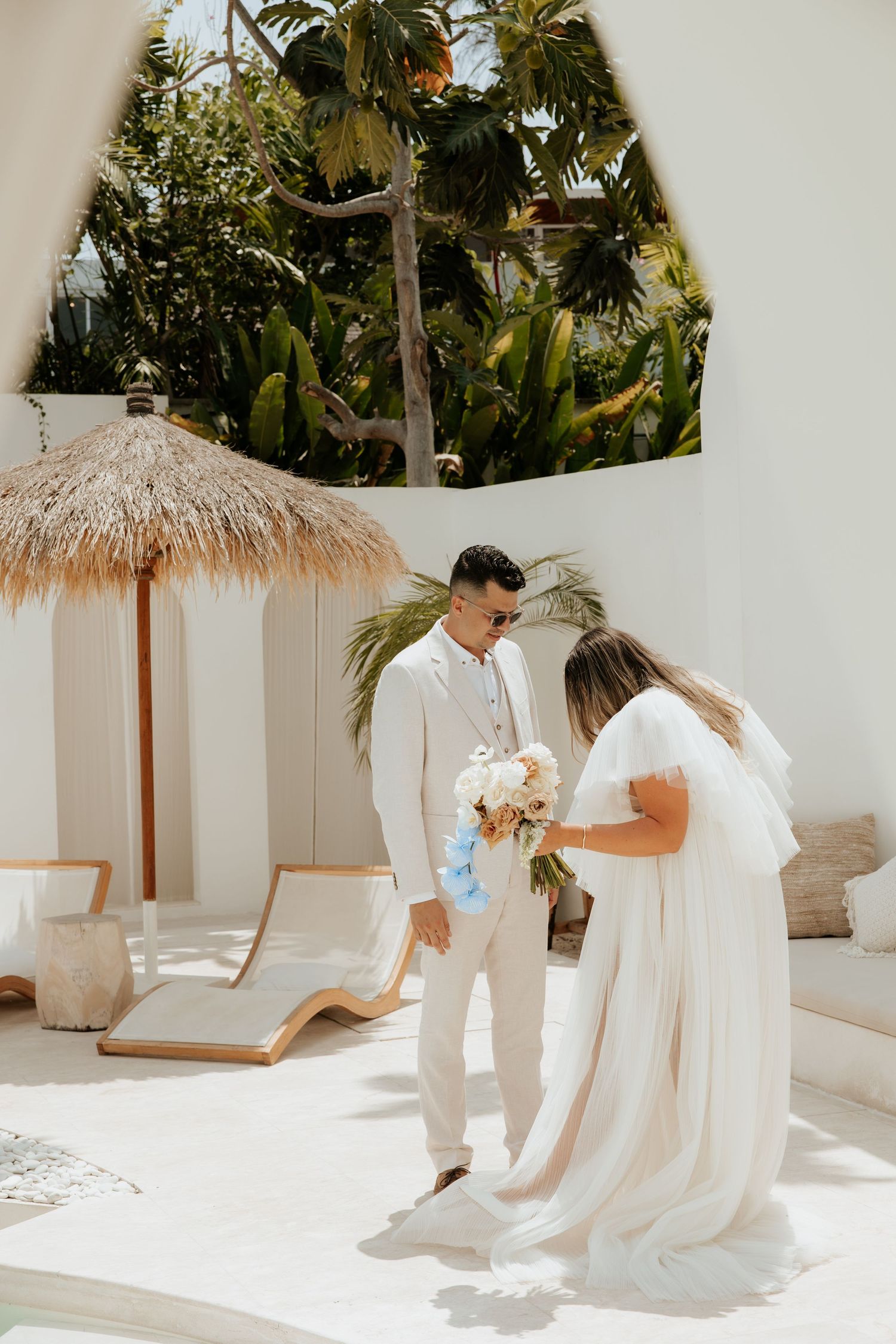 A dreamy wedding scene with palm trees as newlyweds share a moment by a thatched umbrella and lounge chairs.