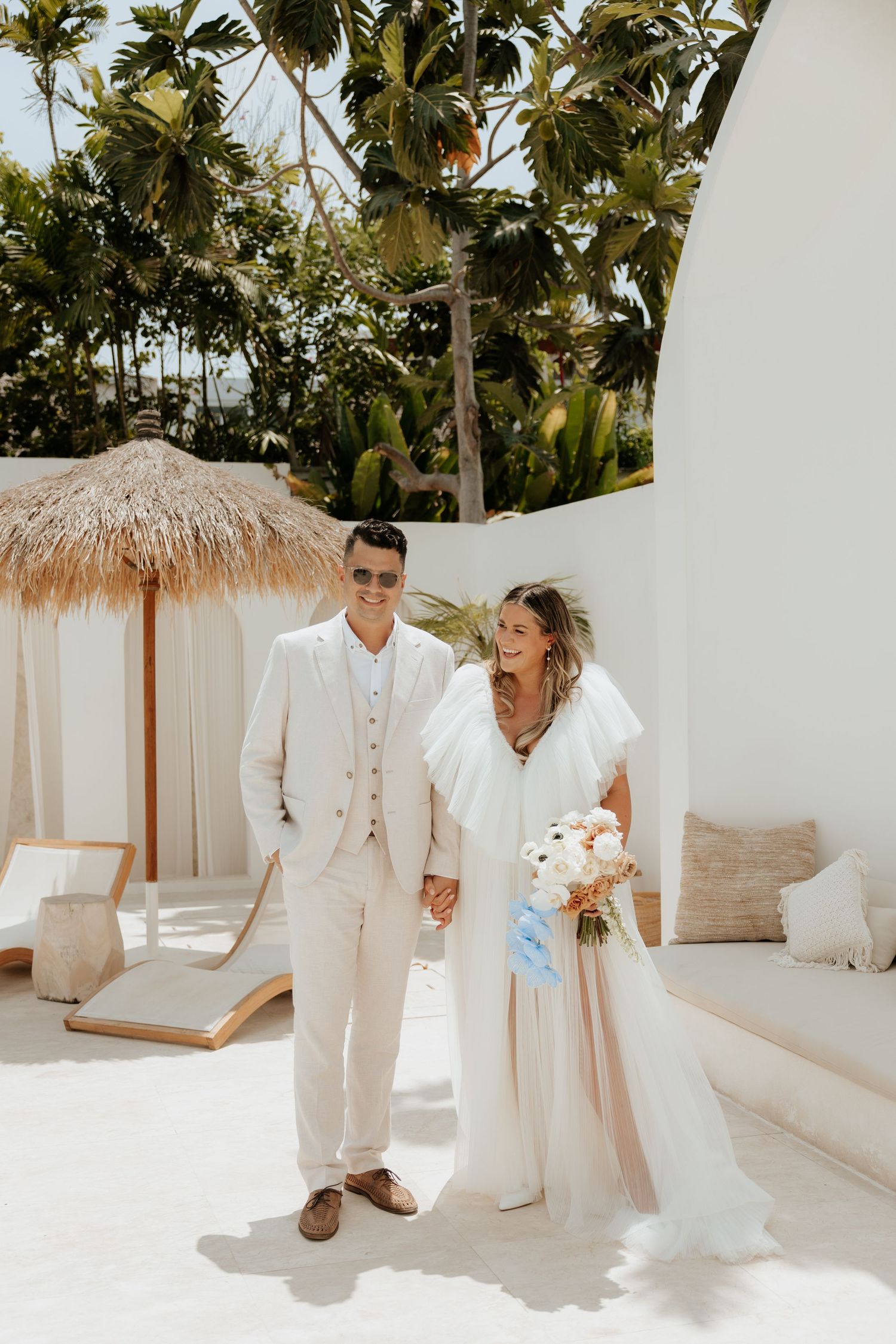 A tropical beach wedding scene with palm trees and a thatched umbrella on a white terrace.