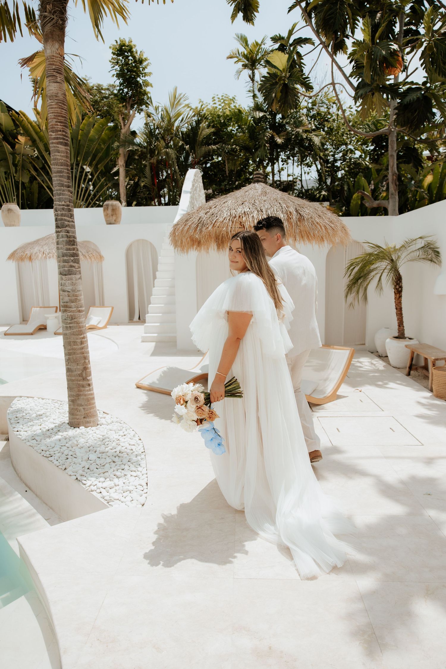 A couple in wedding attire embrace under a thatched umbrella surrounded by palm trees and white decor.