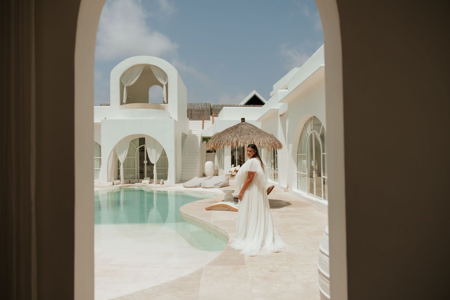 View through arched doorway of Mediterranean style resort with white buildings and turquoise pool surrounded by colonnade.