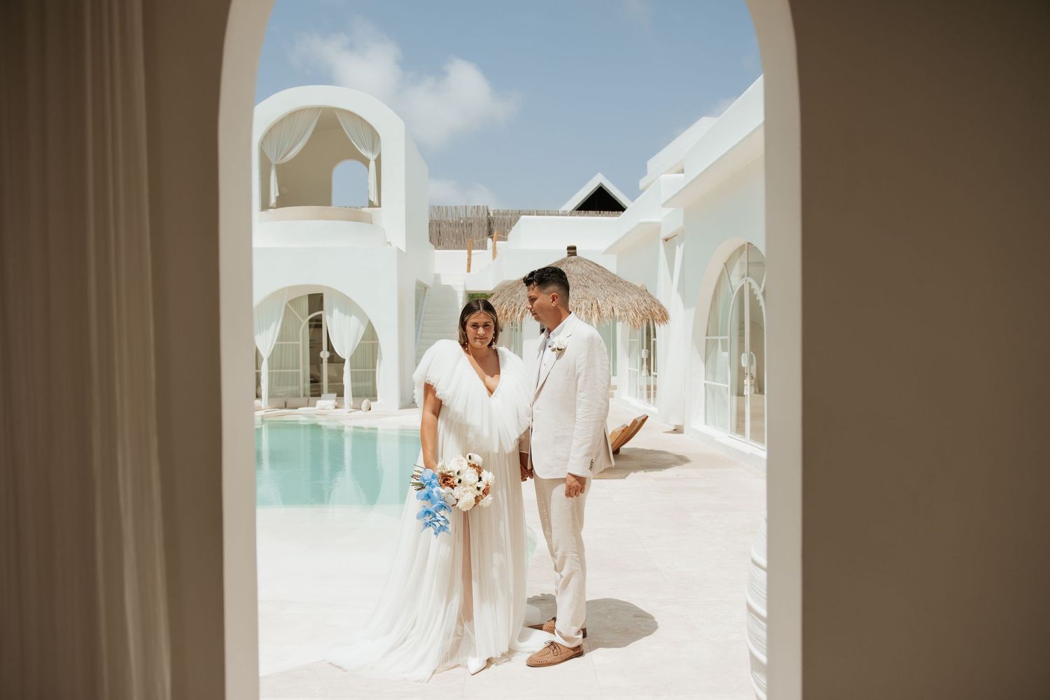 A couple in white outfits stands under an archway at a Mediterranean-style venue with a pool.