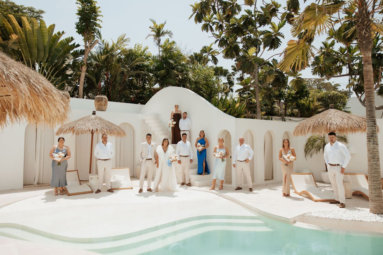 A tropical wedding party poses by a pool with white adobe walls and thatched umbrellas.