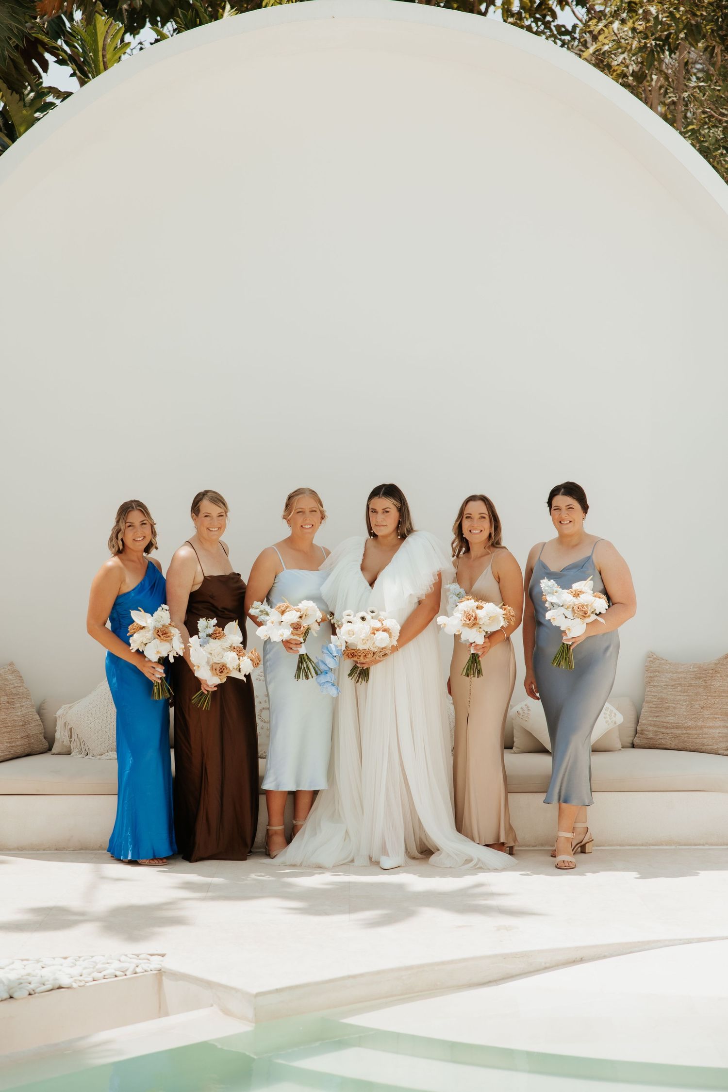 Wedding party posing against a white curved wall by a pool with coordinated pastel-colored dresses.