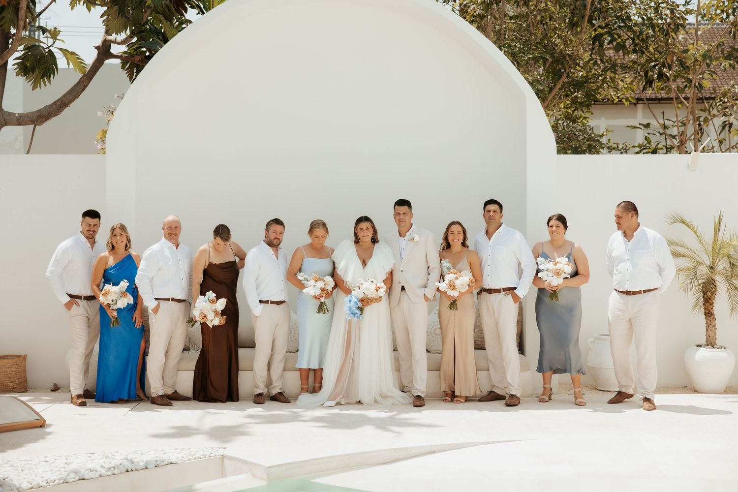 Wedding party members lined up against a white wall in neutral-toned attire for a group photo.