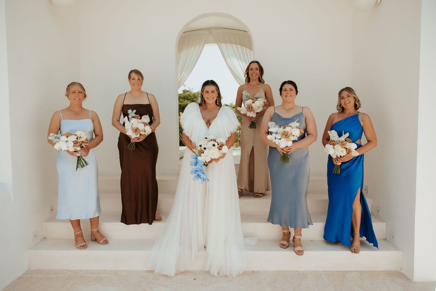 Wedding party standing in white archway wearing coordinated dresses in different colors.