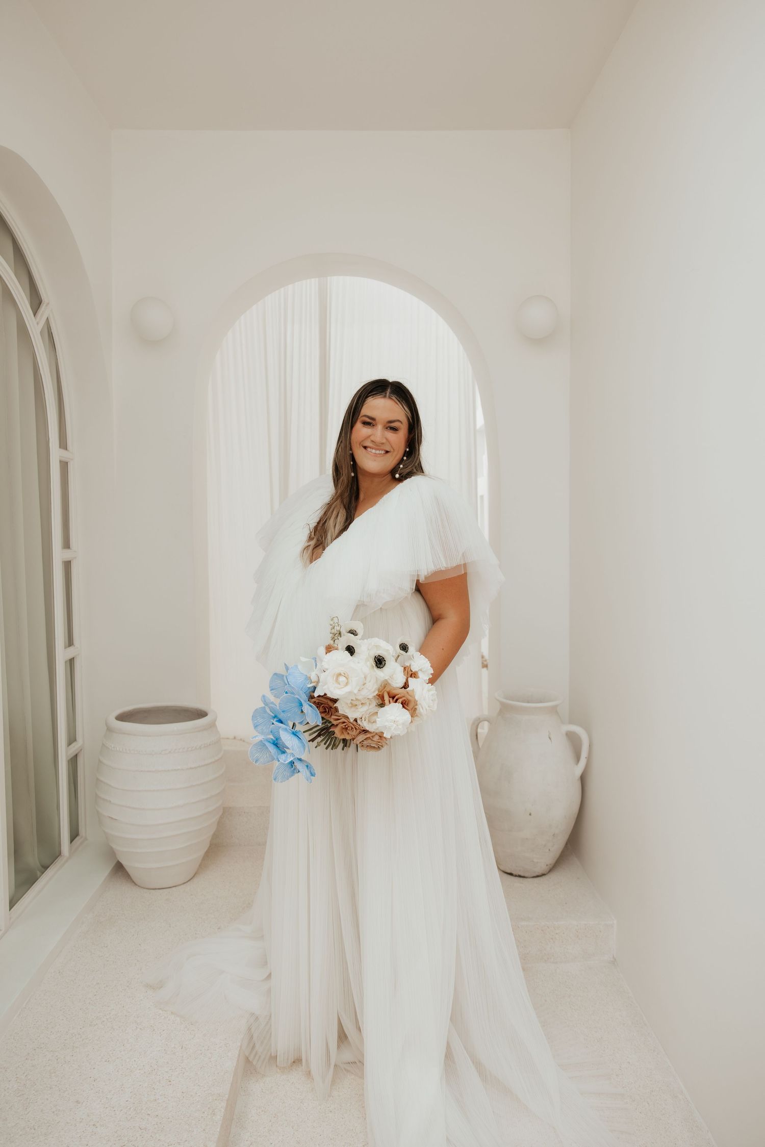 A bride in a white flowing dress holds a blue and white bouquet while standing in an arched white interior space.