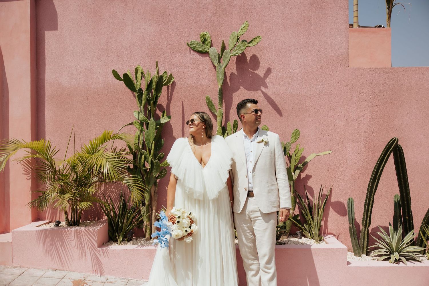 A bride and groom in light colored attire pose against a pink wall with tropical plants and desert landscaping.