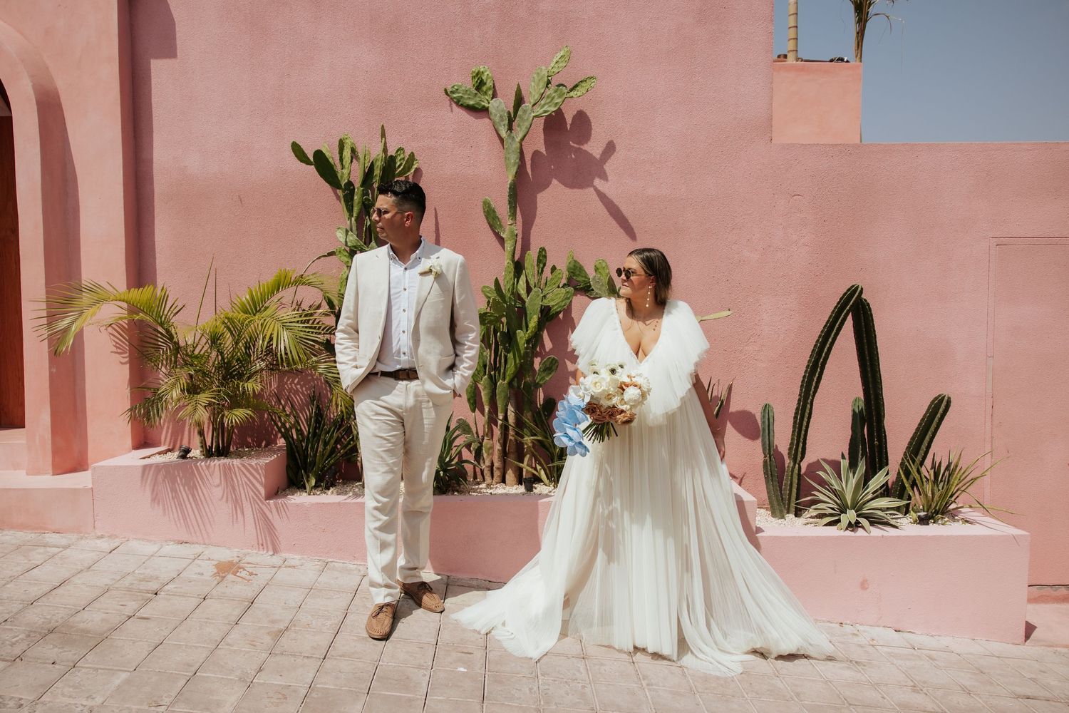 A wedding couple poses against a pink wall with tropical plants at a destination ceremony.
