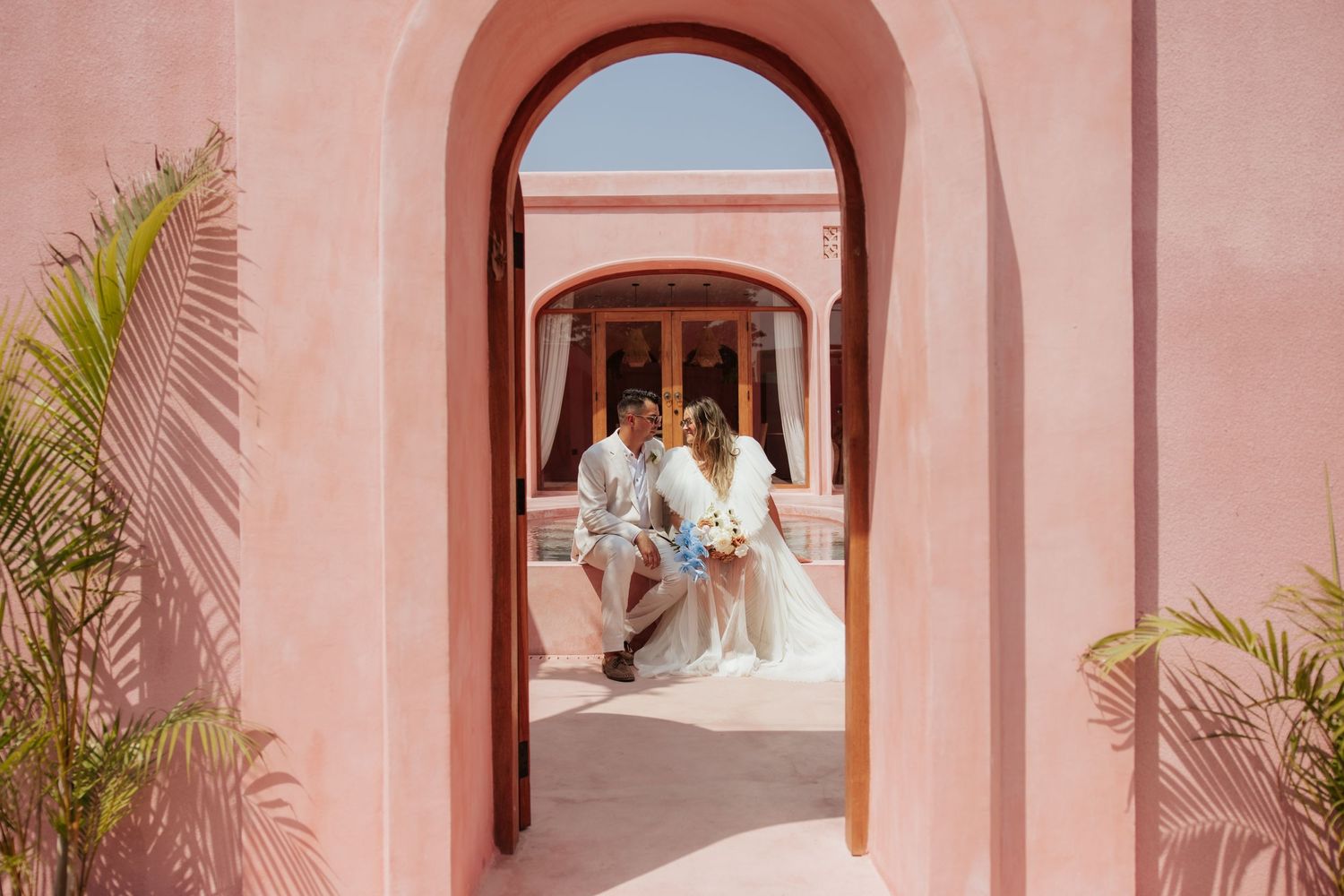 An elegant wedding photo framed through a pink archway with palm trees in the background.