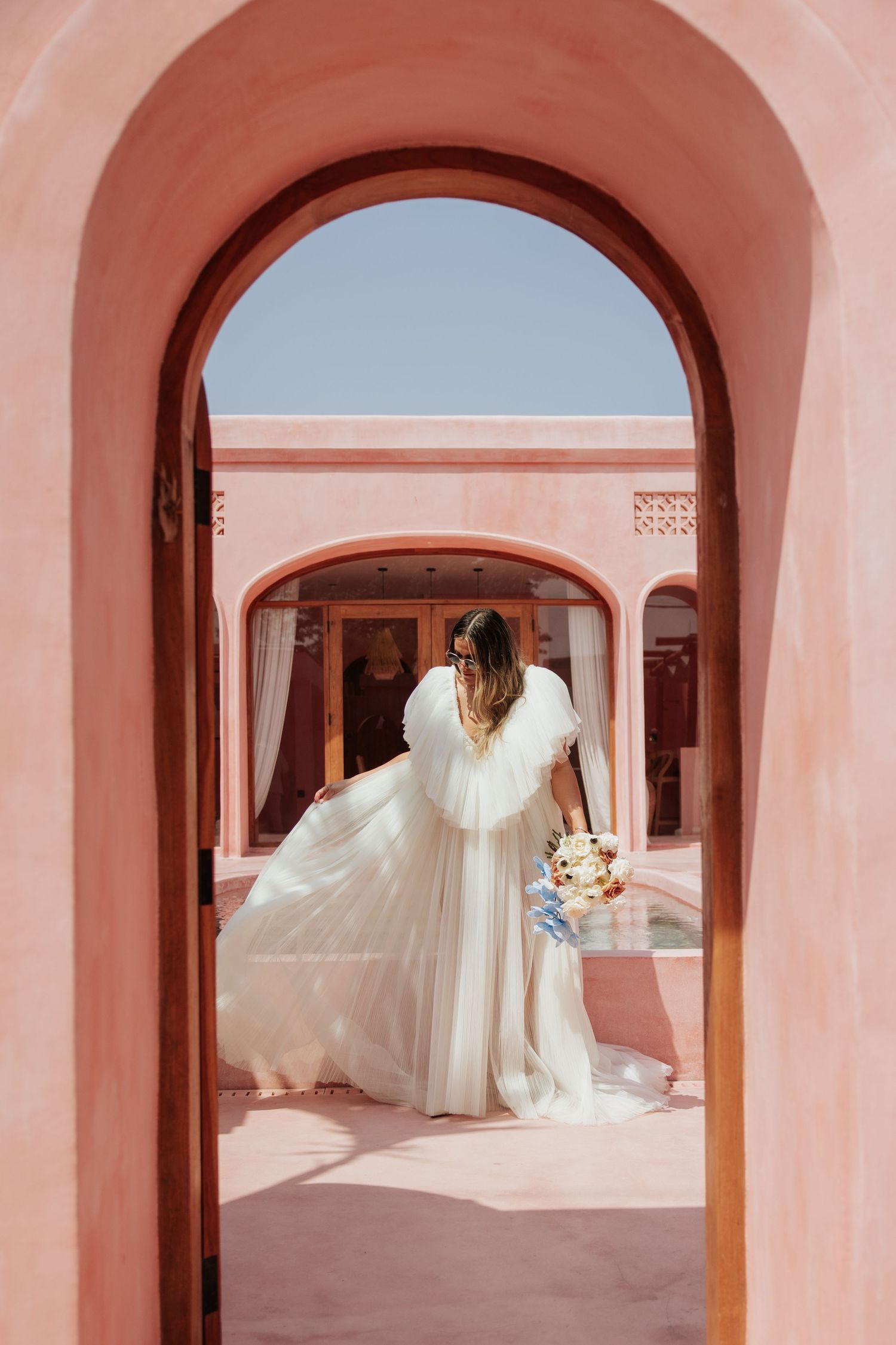 A bride in a flowing white dress and veil poses under a pink archway in an elegant courtyard setting.