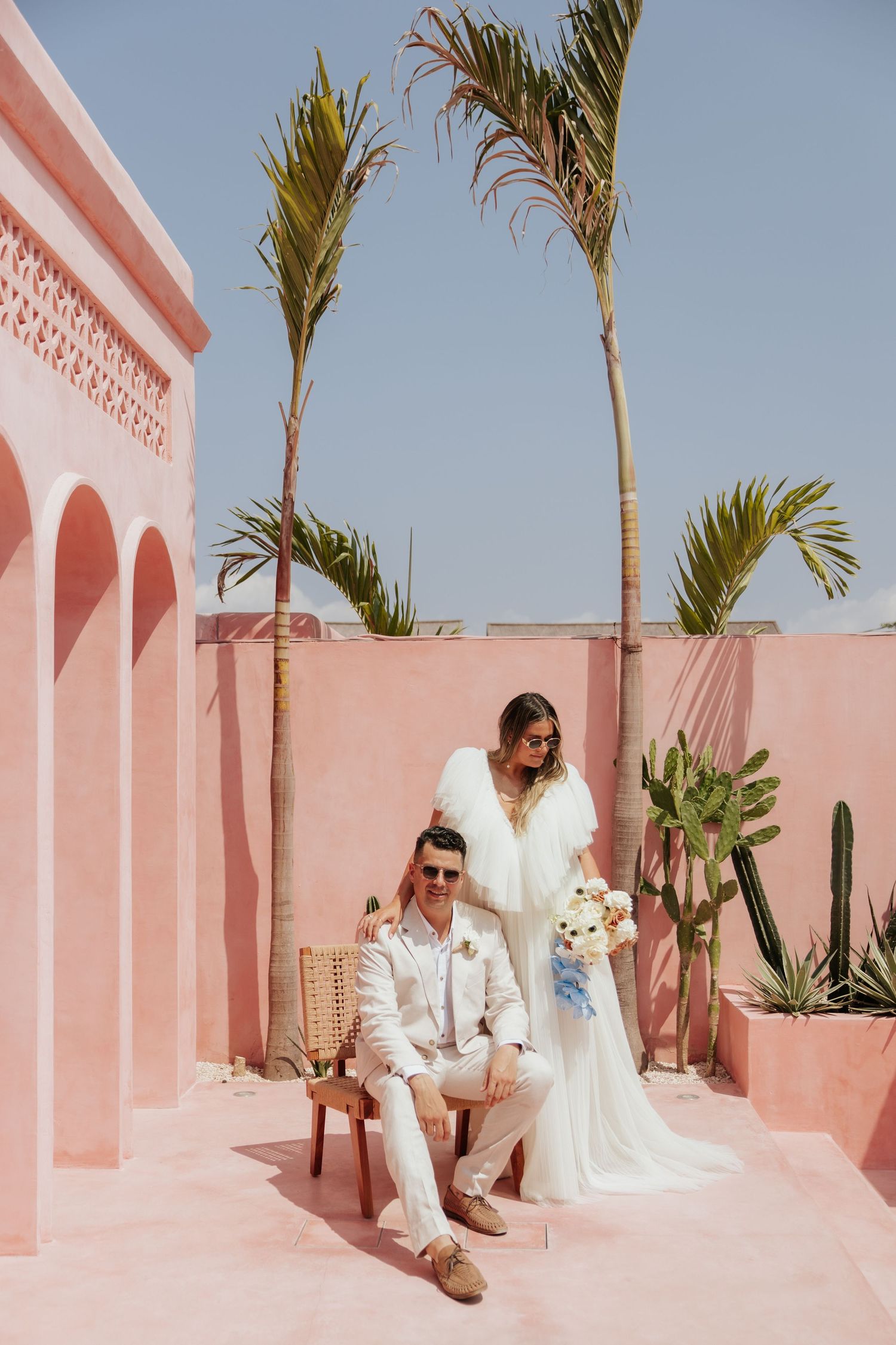 Wedding couple posing in front of pink walls and palm trees at a tropical venue.