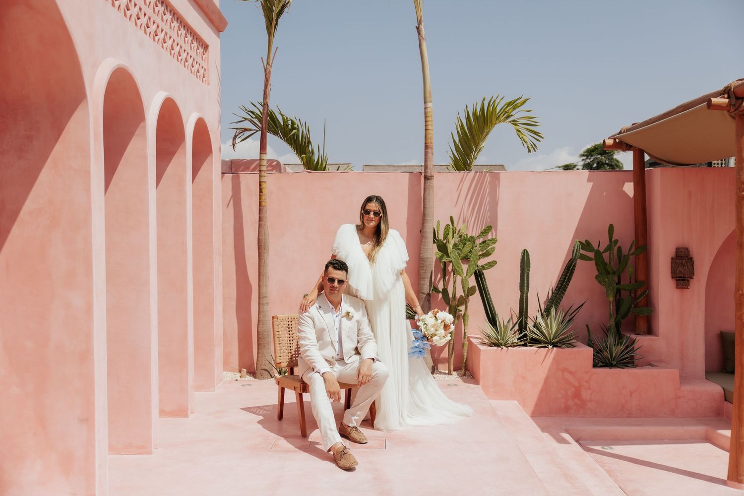 Couple in wedding attire sitting on pink stairs with palm trees and Mediterranean architecture.