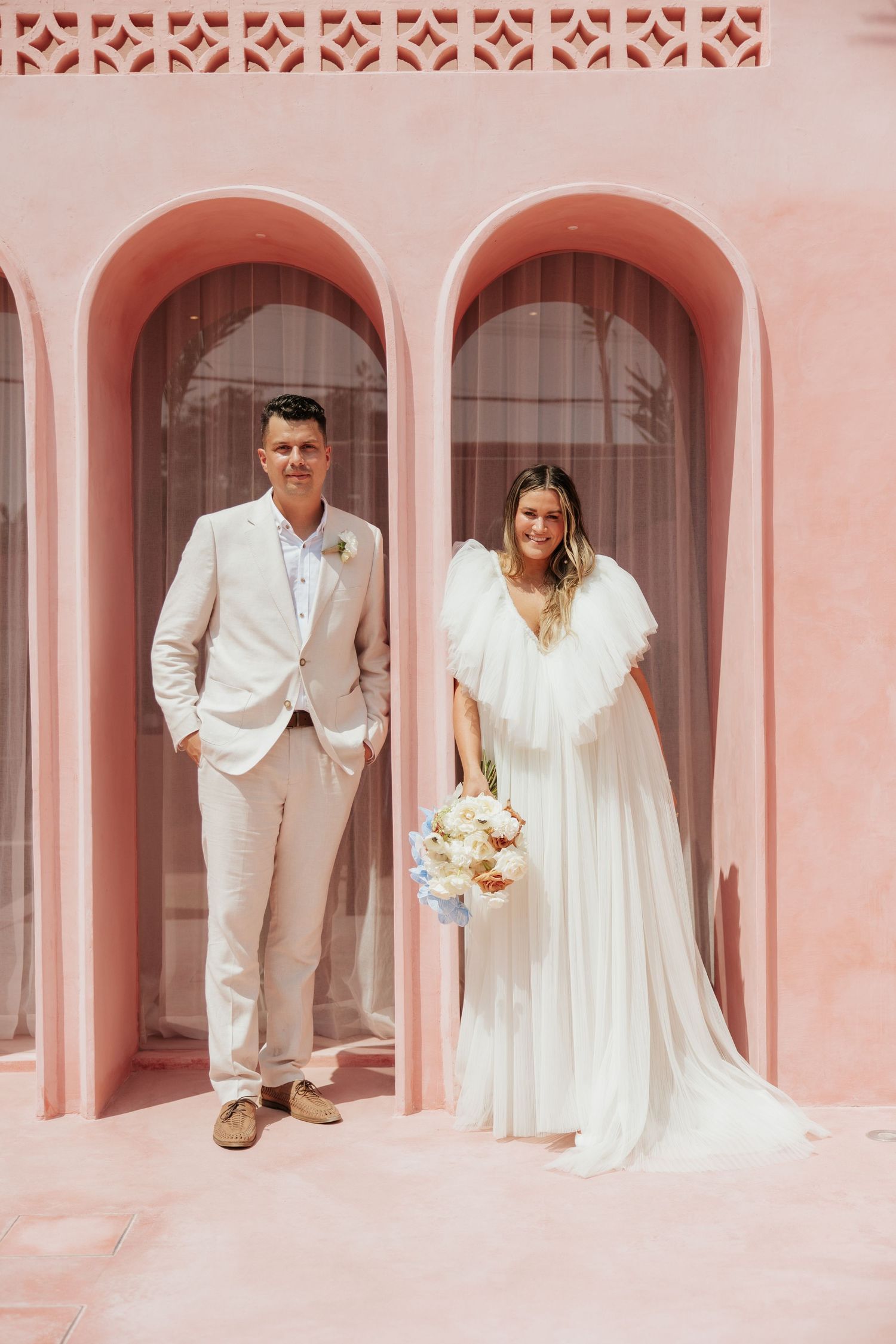 A wedding couple poses in front of pink arched doorways in matching light-colored attire.