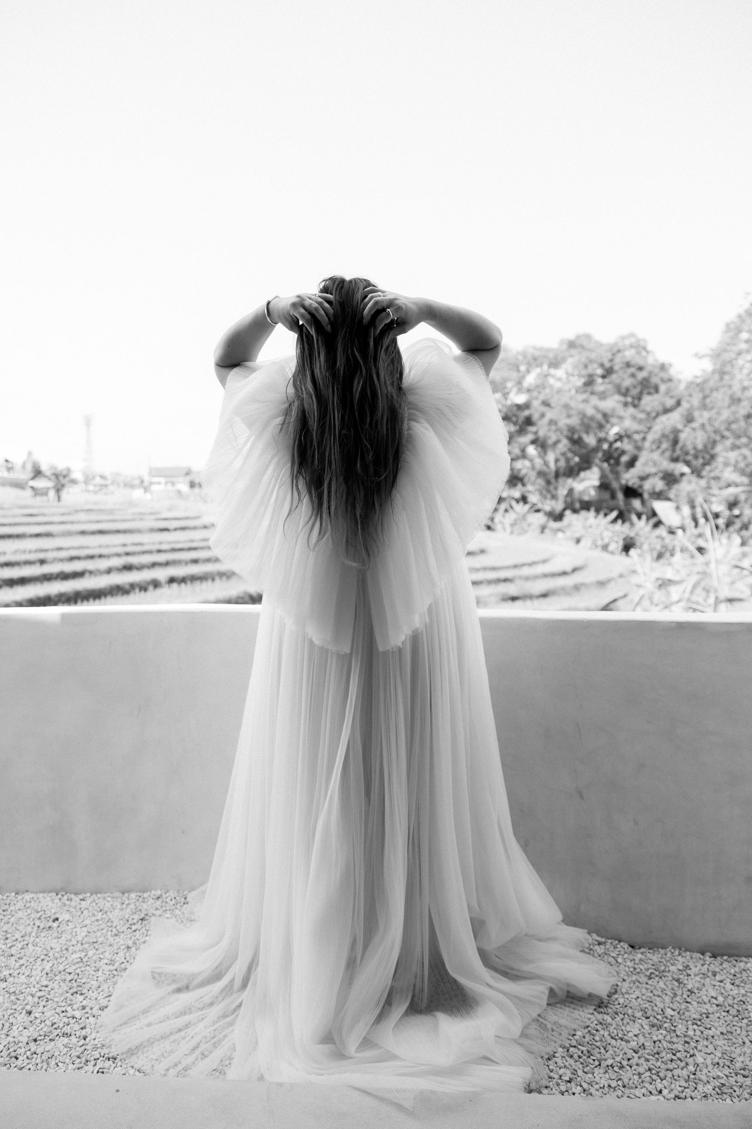 Black and white photo of bride in flowing wedding dress shown from behind outdoors.