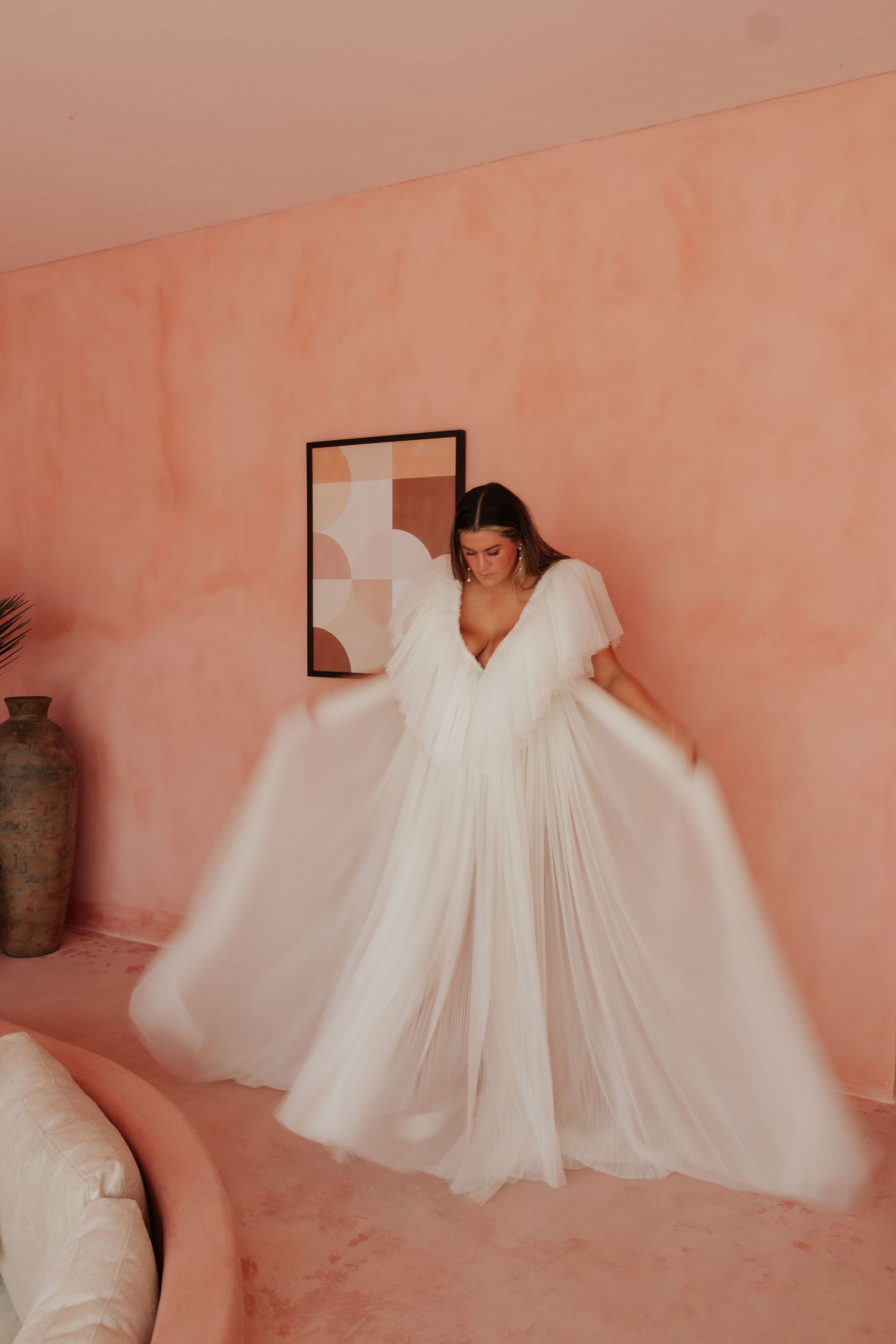 A flowing white wedding dress with dramatic tulle sleeves is displayed against a peachy pink wall with minimal decor.