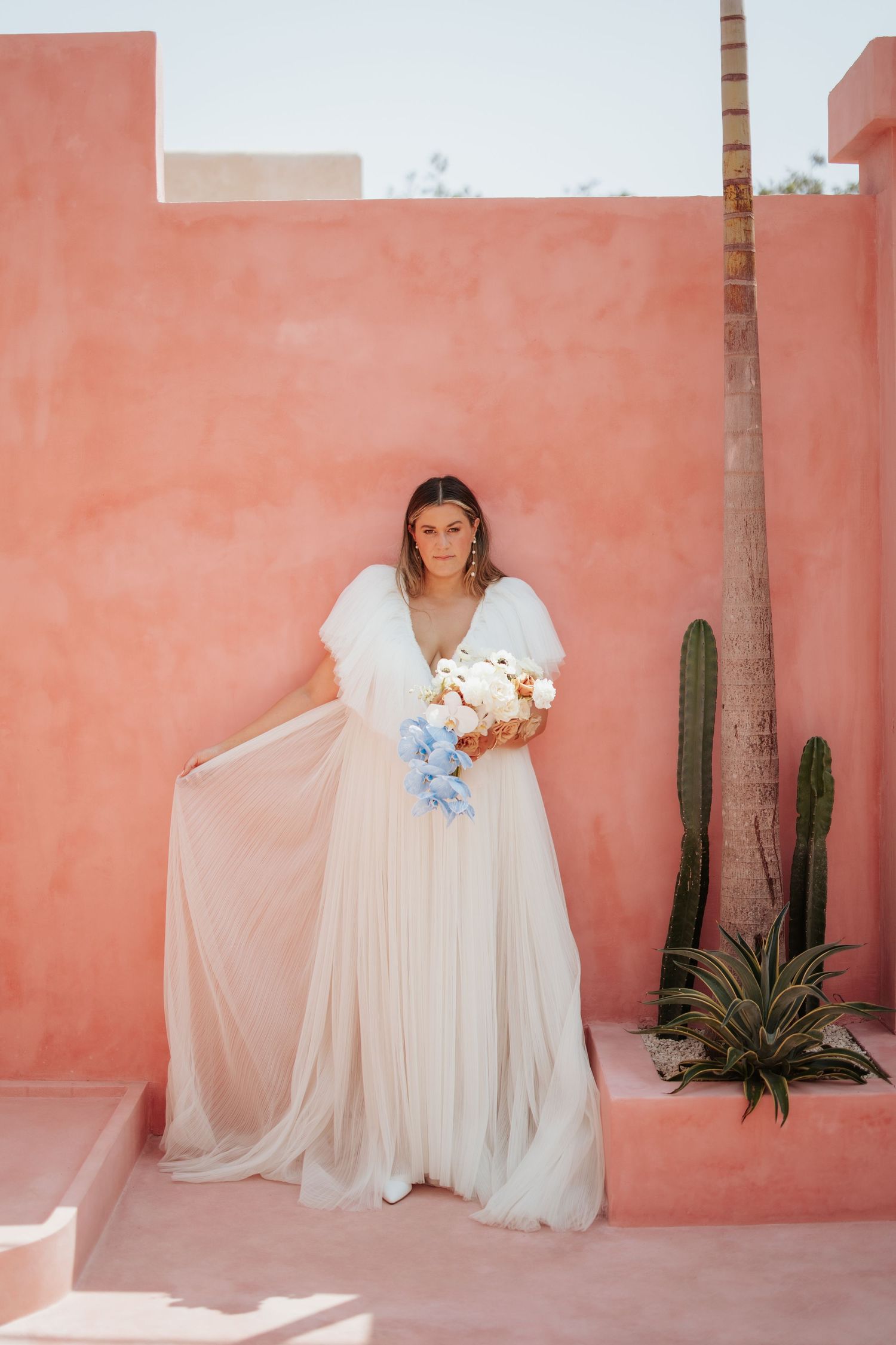 A bride in a flowing white dress holds a bouquet against a coral-colored wall with desert landscaping and palm trees.