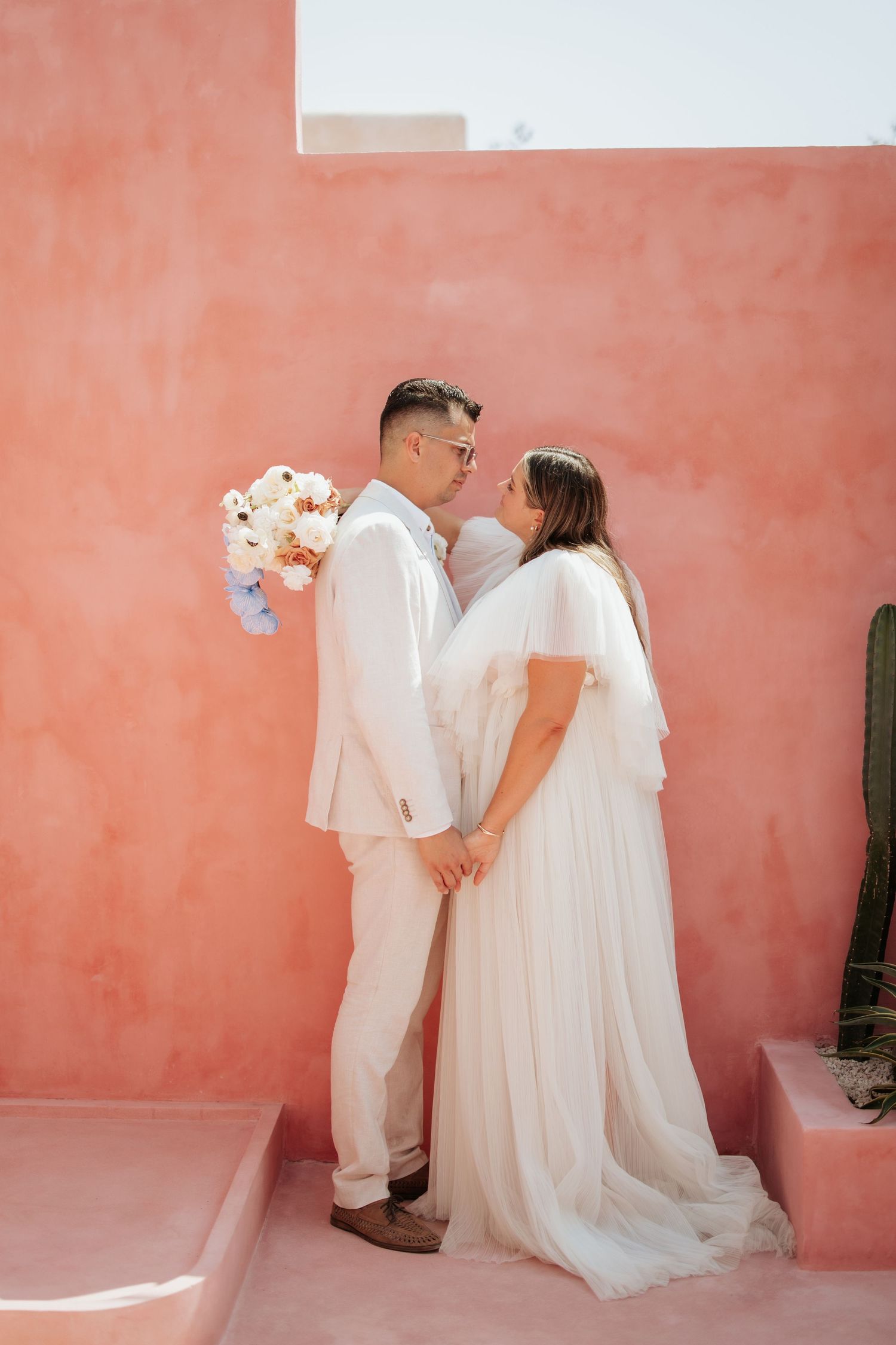 Newlyweds in white attire share an intimate moment against a coral-colored wall in a romantic wedding portrait.