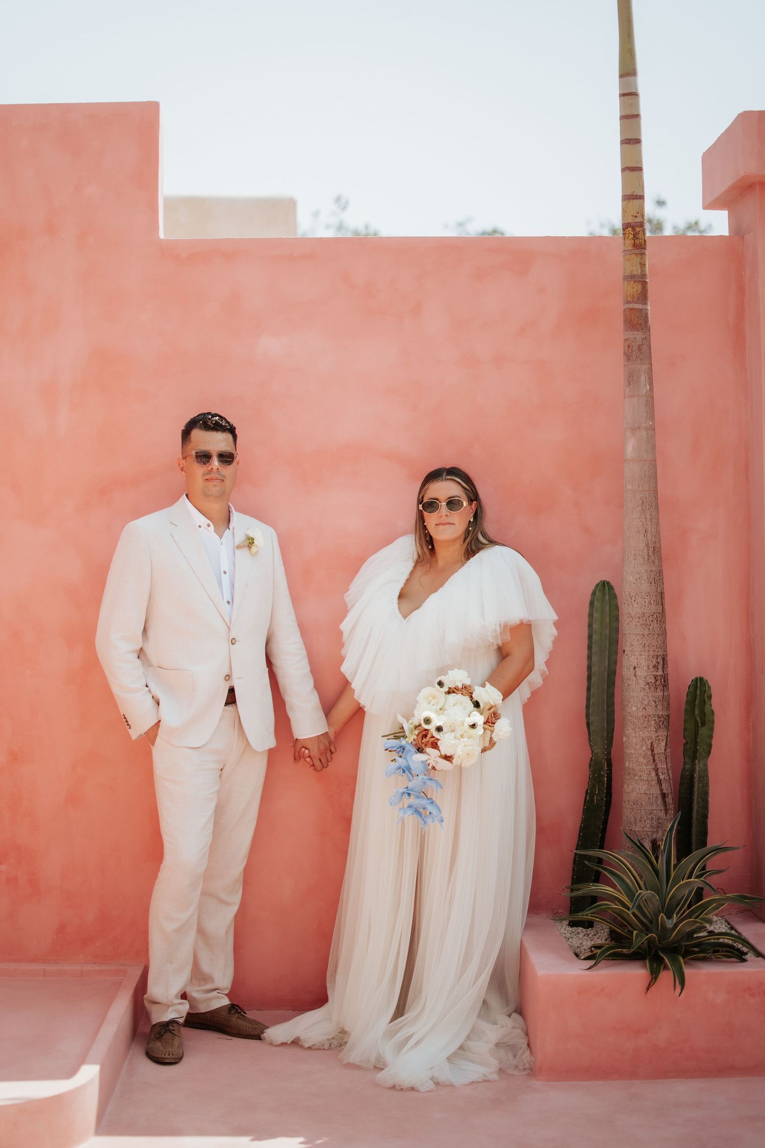 A couple in light colored wedding attire poses against a coral-colored wall with a cactus and palm tree in the background.