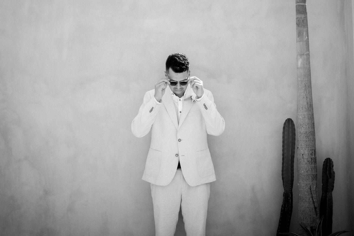 A black and white photo of a person in a white suit and sunglasses against a neutral wall.