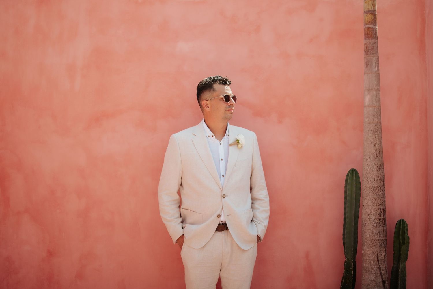 Someone in a light colored suit and sunglasses poses against a pink wall with a palm tree and cactus.