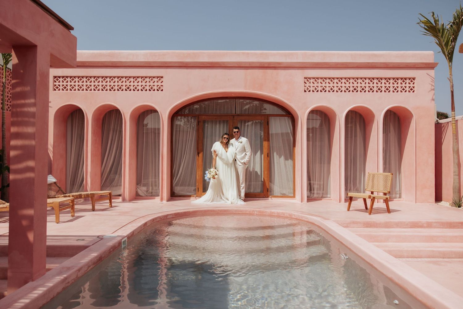 Two people embrace at a luxurious pink Mediterranean-style villa with arched windows and a reflecting pool.