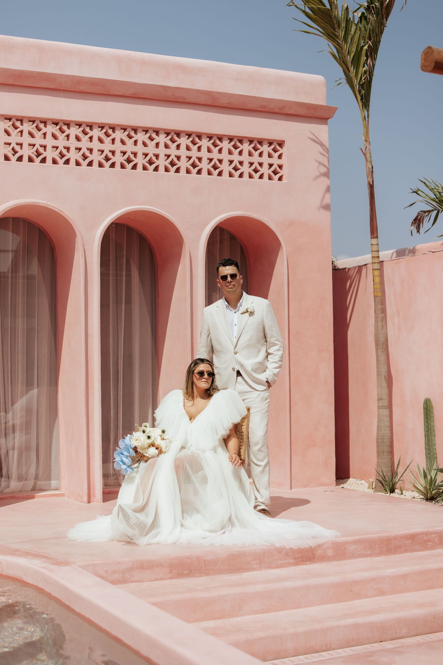 A wedding couple sits on pink steps of a Mediterranean-style building with arches and palm trees.