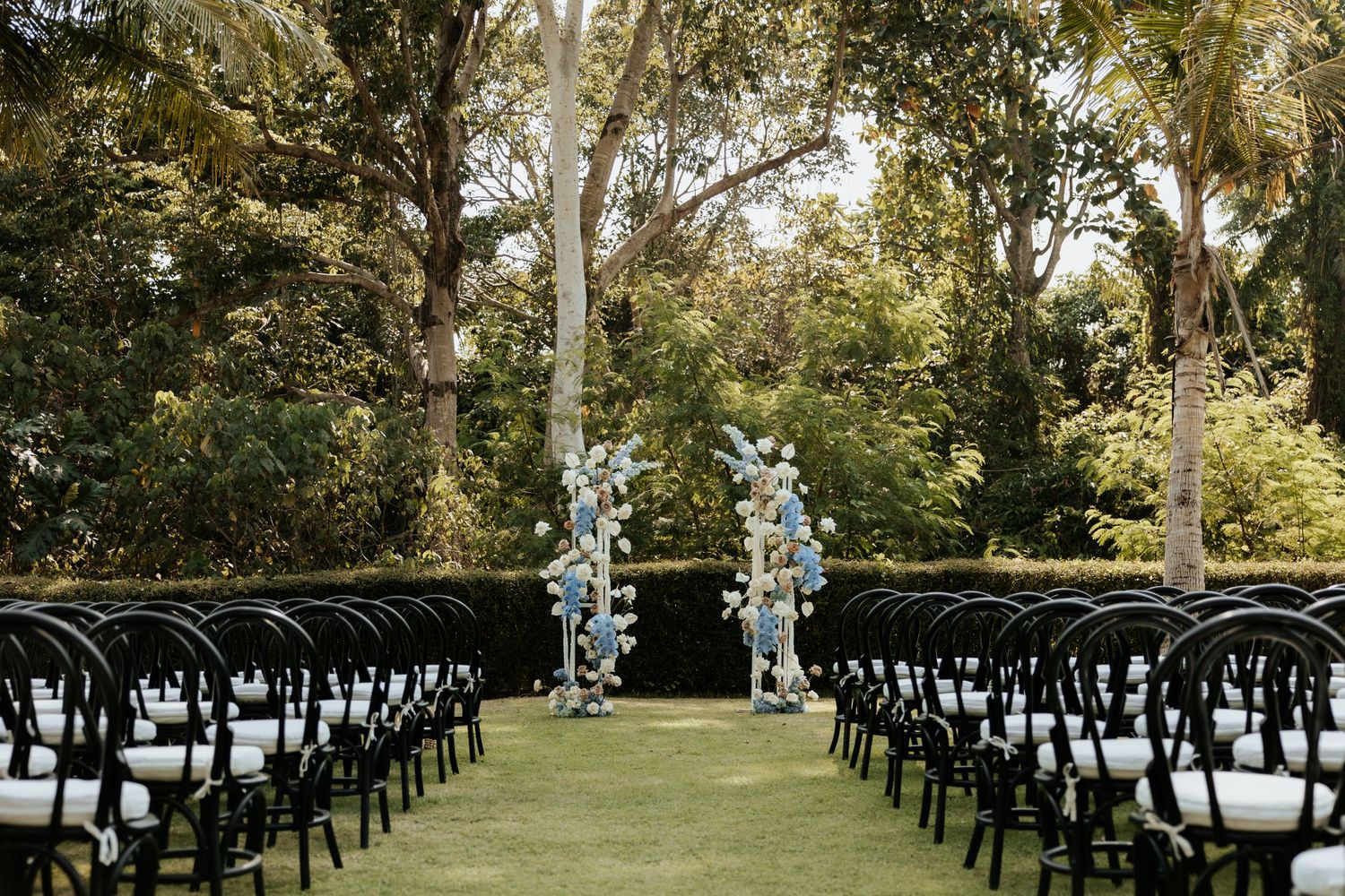 Outdoor wedding ceremony setup with black chairs and floral arch in a garden setting.