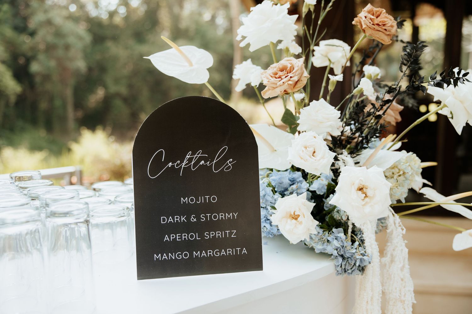 Elegant black cocktail menu sign with white lettering displayed next to delicate white and blush floral arrangements.