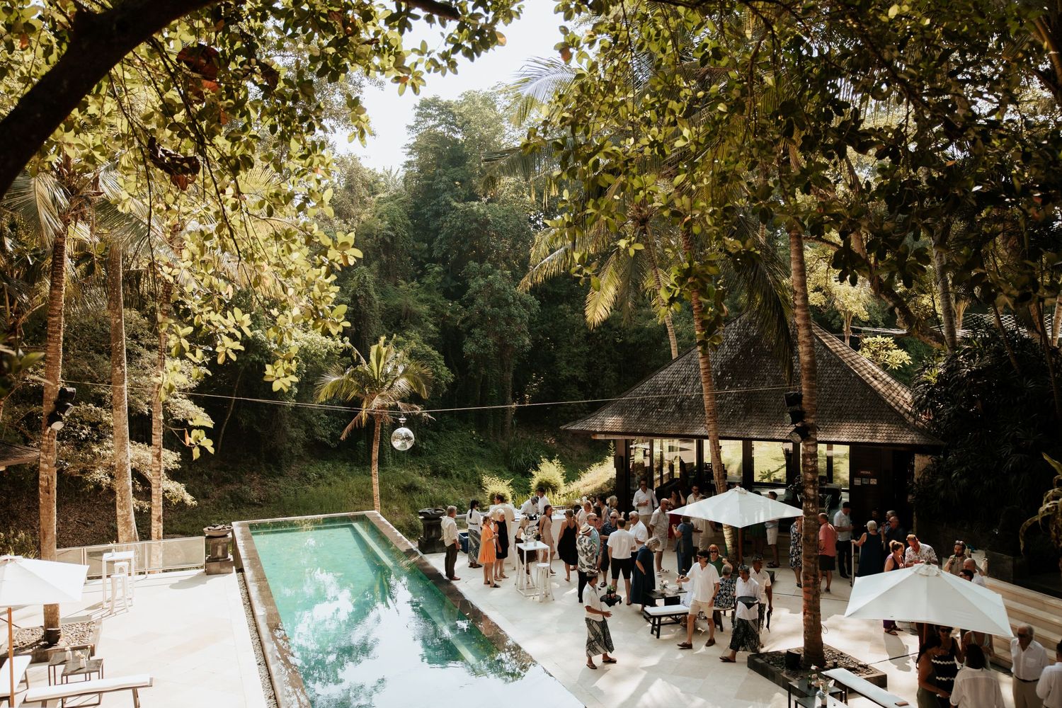 Outdoor wedding reception by a swimming pool surrounded by lush trees and umbrellas in a tropical setting.
