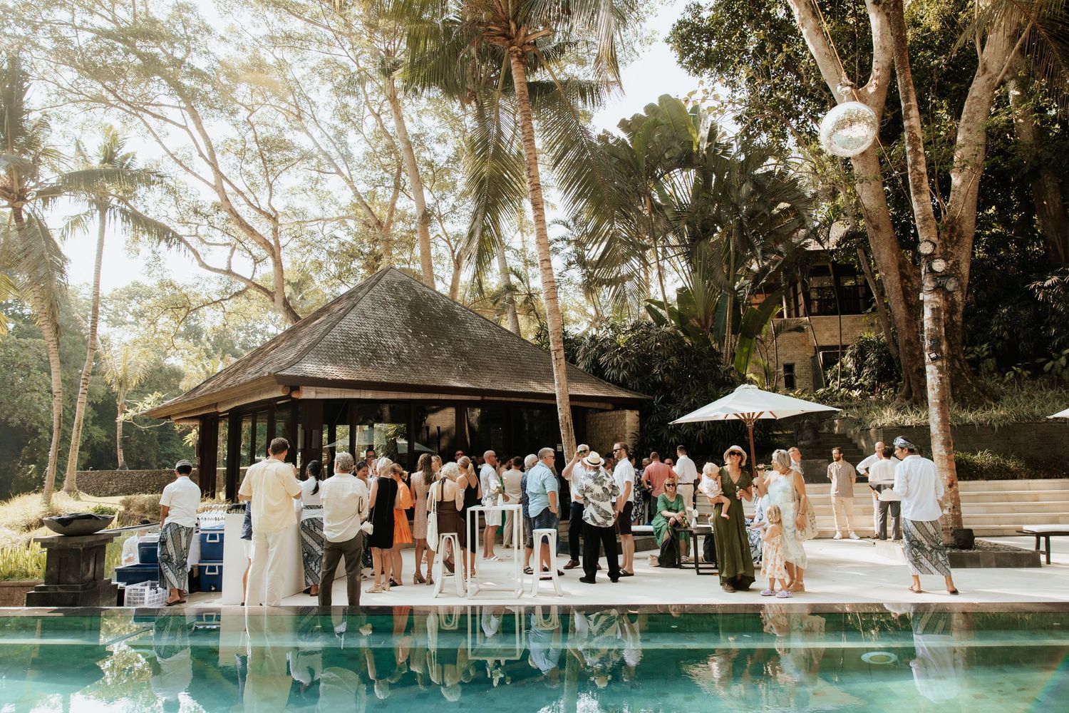 Tropical pool party gathering at a thatched-roof pavilion surrounded by lush palm trees.