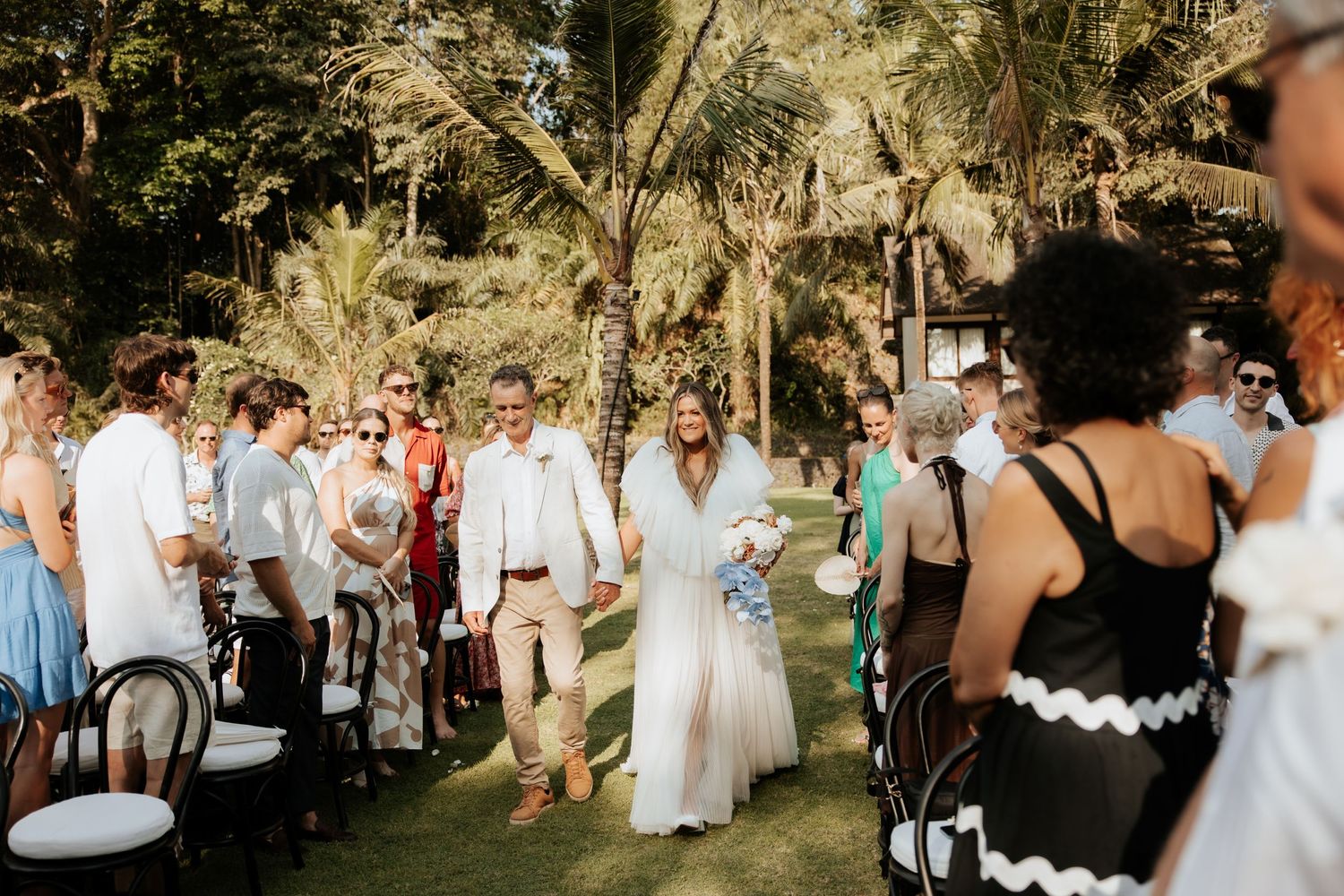Wedding guests stand during an outdoor ceremony surrounded by lush tropical greenery.