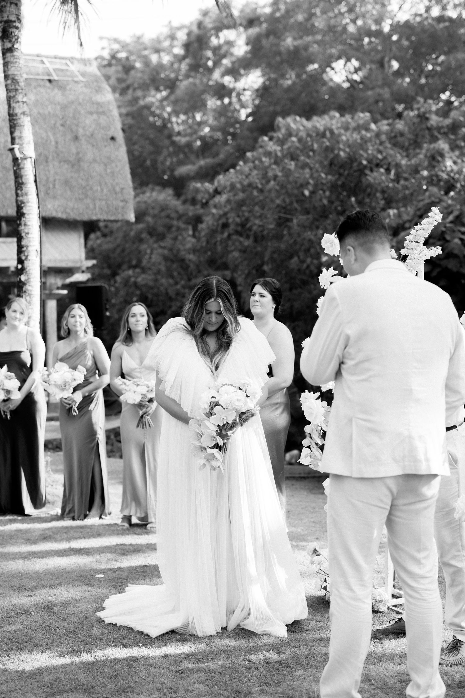 A black and white outdoor wedding ceremony with bridesmaids in flowy dresses standing in line behind the bride.