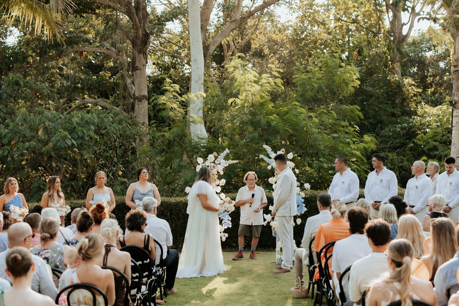 An outdoor wedding ceremony in a lush garden setting with guests seated on white chairs.