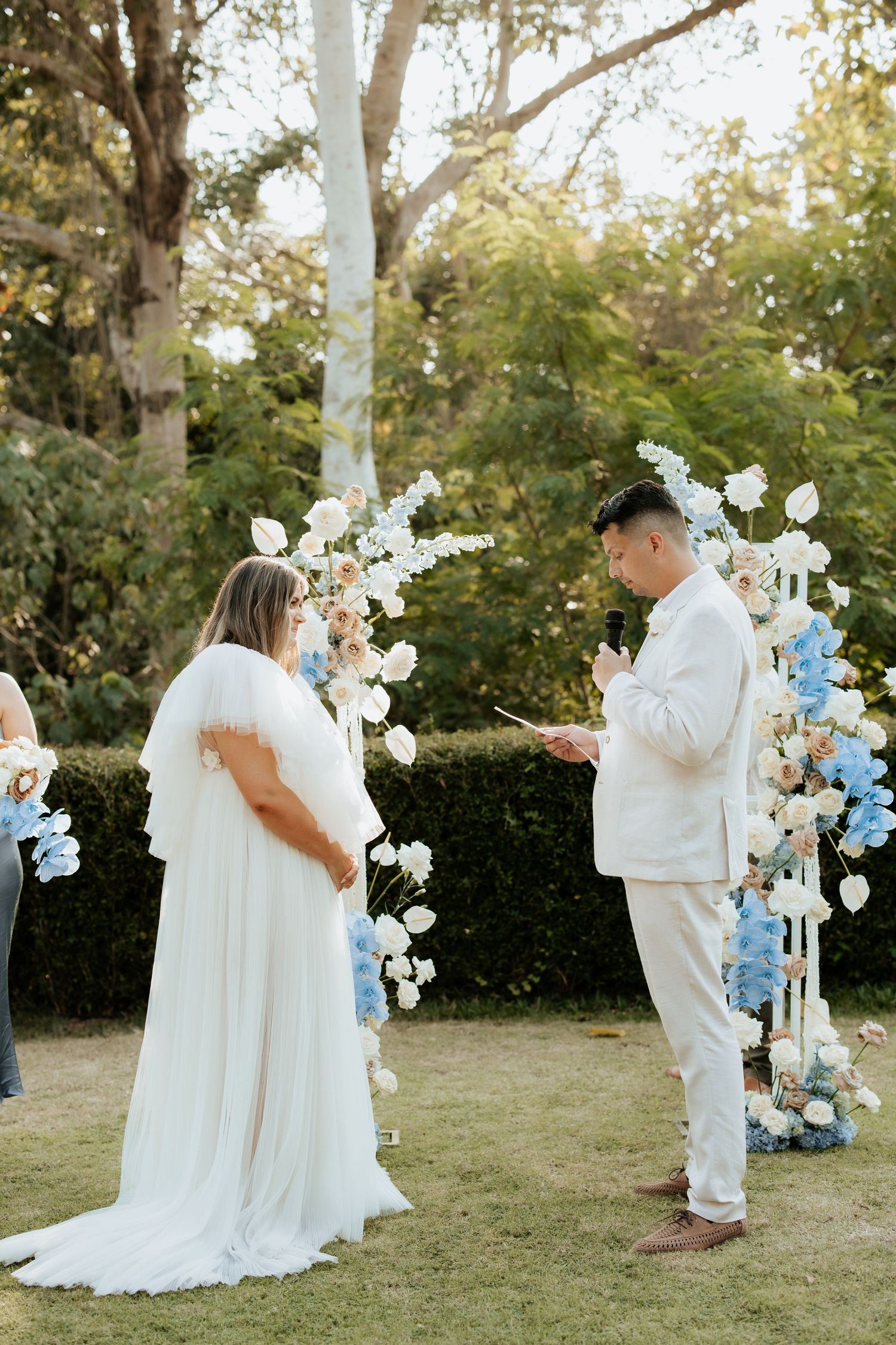 A wedding ceremony under a floral arch with blue and white flowers in an outdoor garden setting.