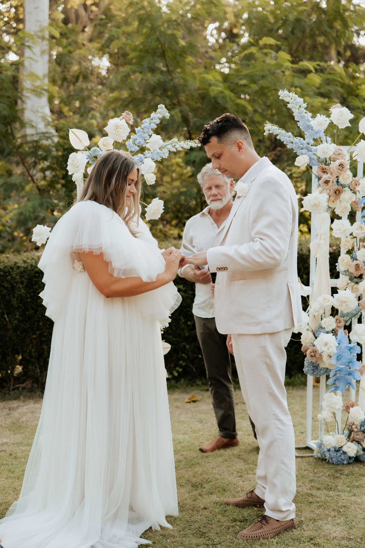 An intimate outdoor wedding ceremony with floral arch decor and participants dressed in white attire.