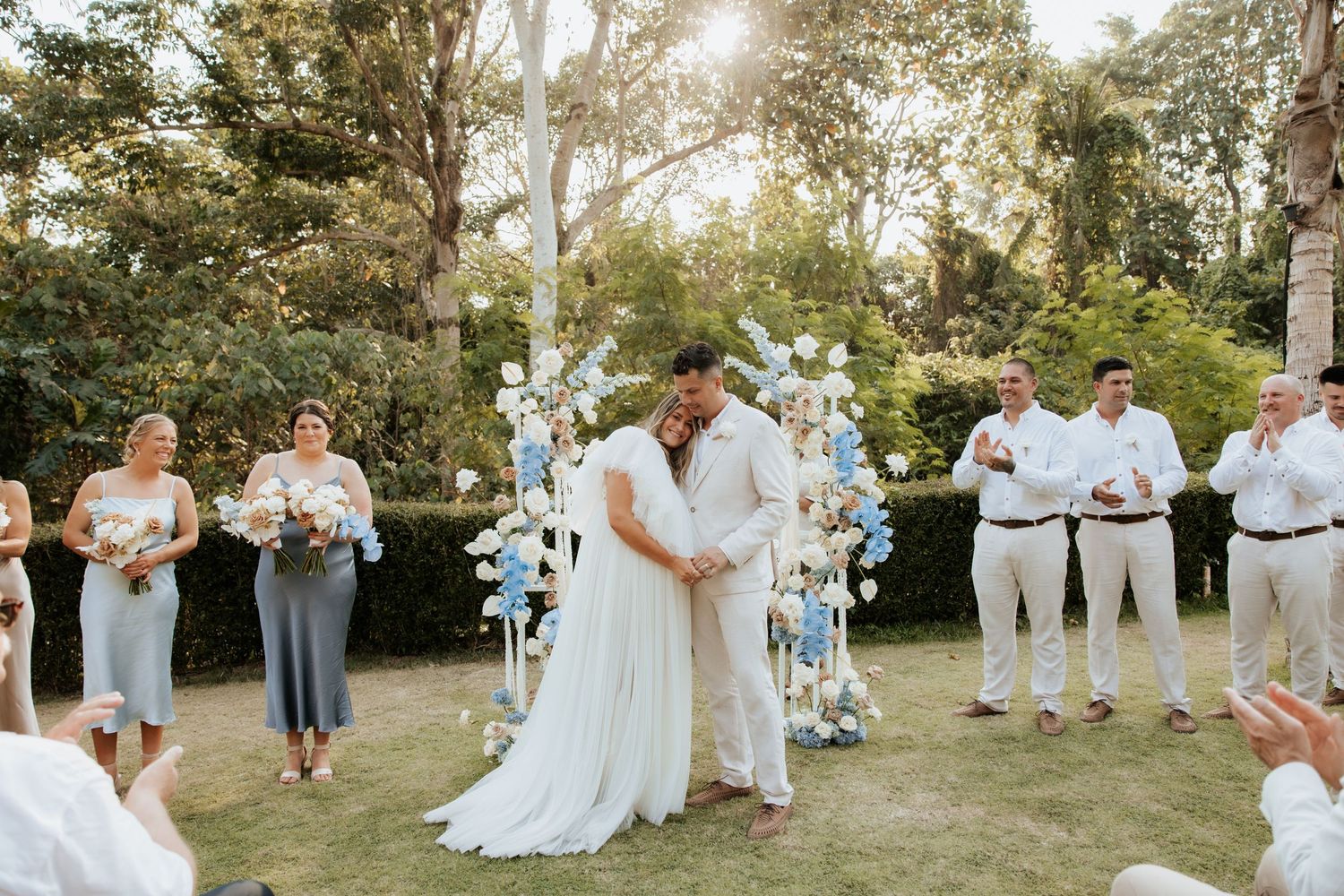 Wedding ceremony taking place outdoors with floral arch decorations and guests watching in a garden setting.