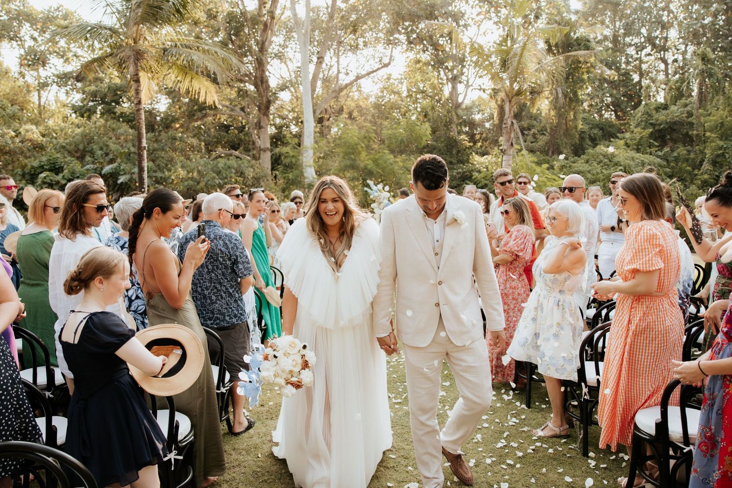 A couple walks down an outdoor aisle lined with guests during an outdoor wedding ceremony.